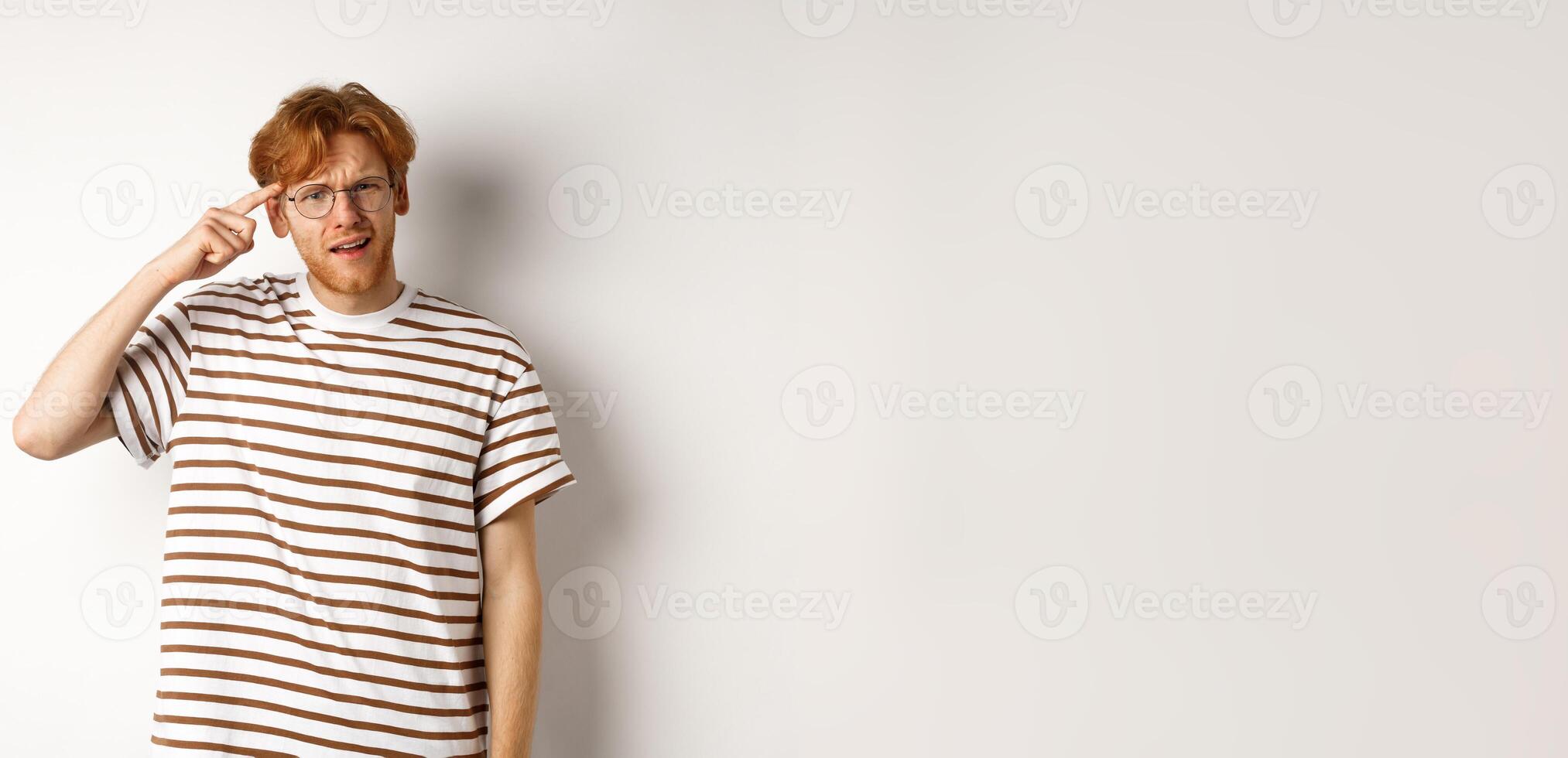 Annoyed young man with red hair scolding person for being stupid, pointing at head and staring at camera, white background photo