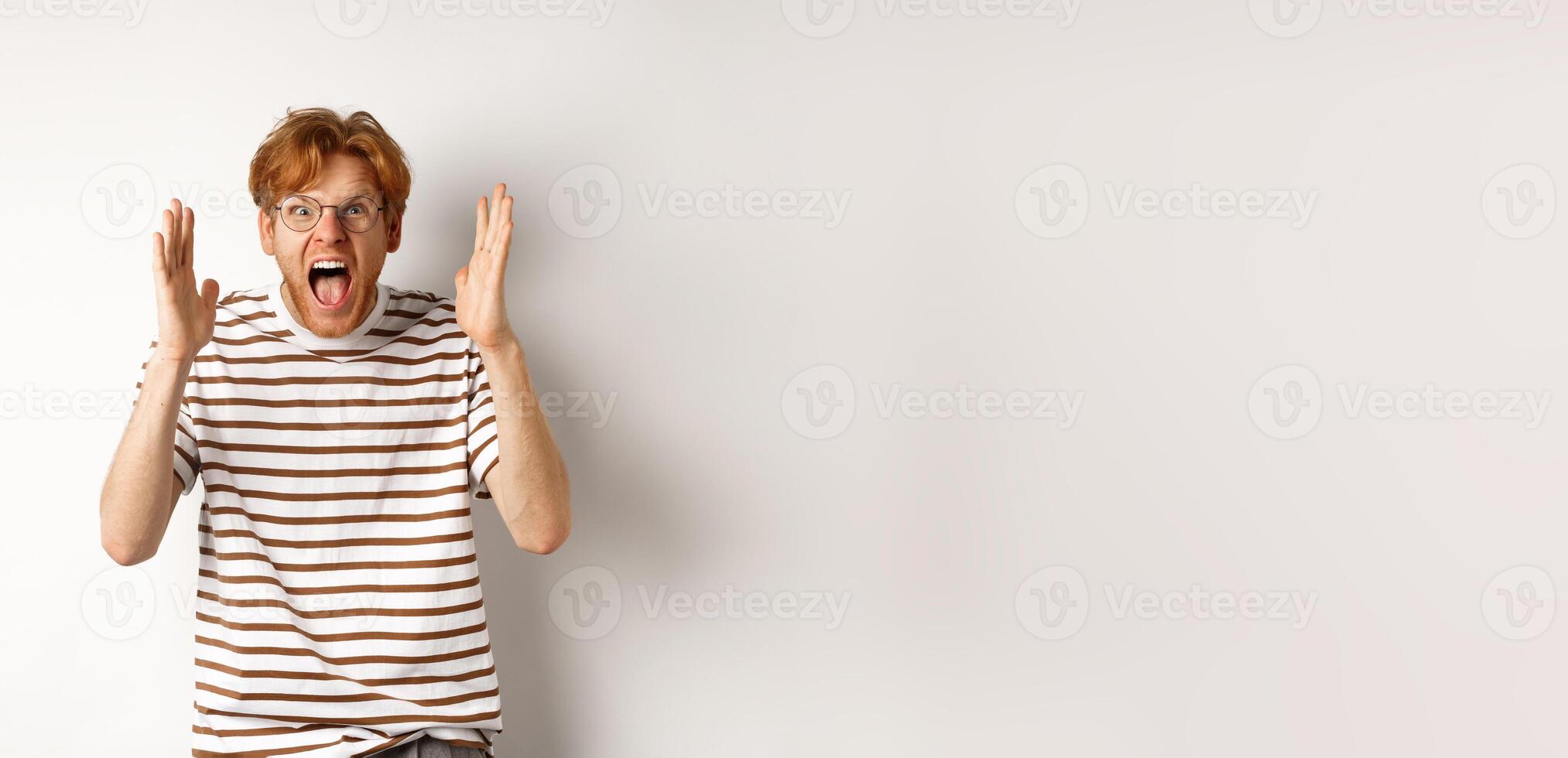 Angry young man with red hair shouting at camera, screaming and looking outraged, shaking hands, standing over white background photo