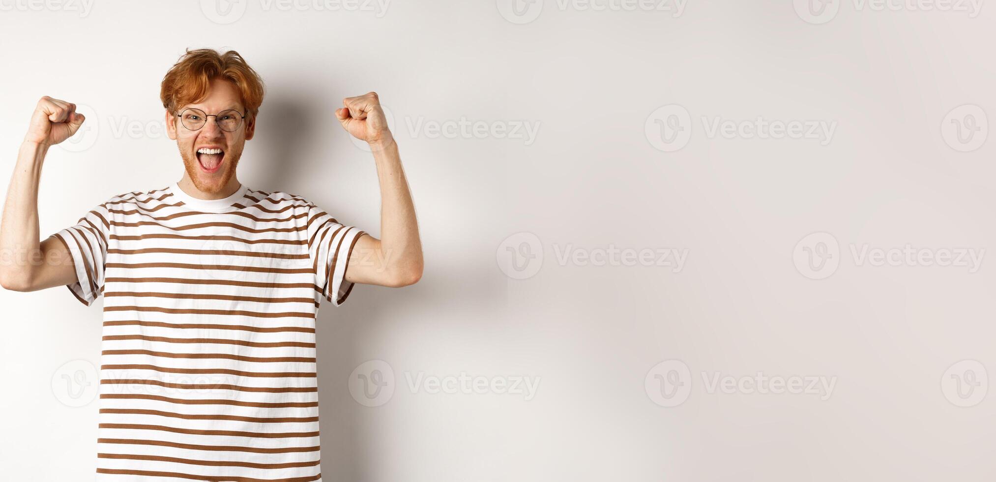 Joyful redhead guy raising hands up like winner, winning prize and celebrating, shouting for joy, standing over white background photo