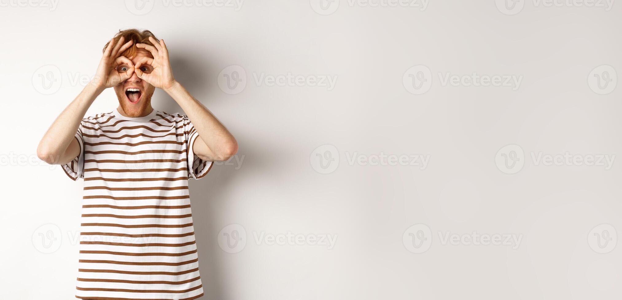 Impressed redhead man checking out promo offer, looking at camera from hand binoculars, smiling amazed, white background photo