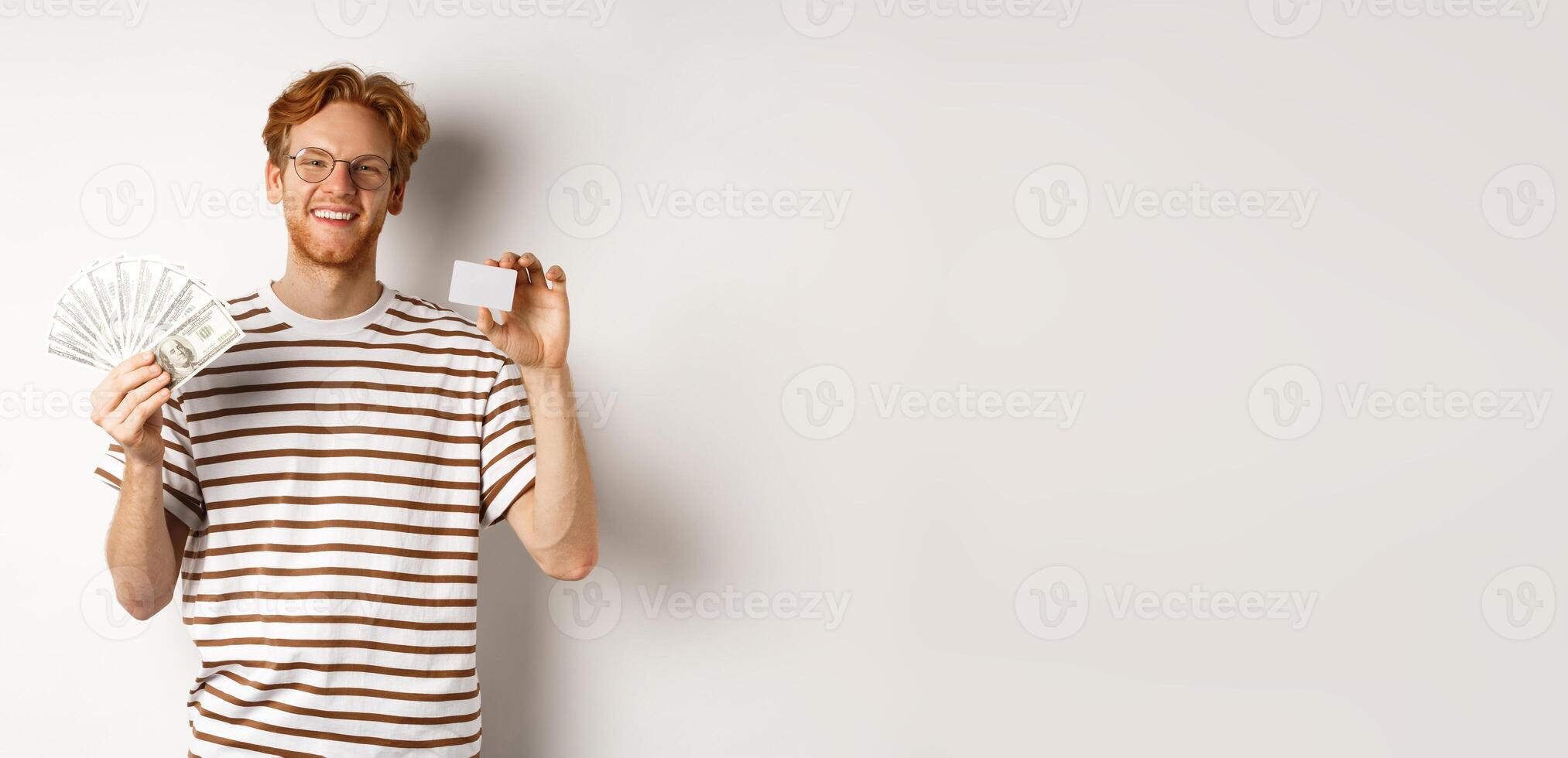 Shopping and finance concept. Young redhead man with beard and glasses showing plastic credit card with money in dollars, white background photo