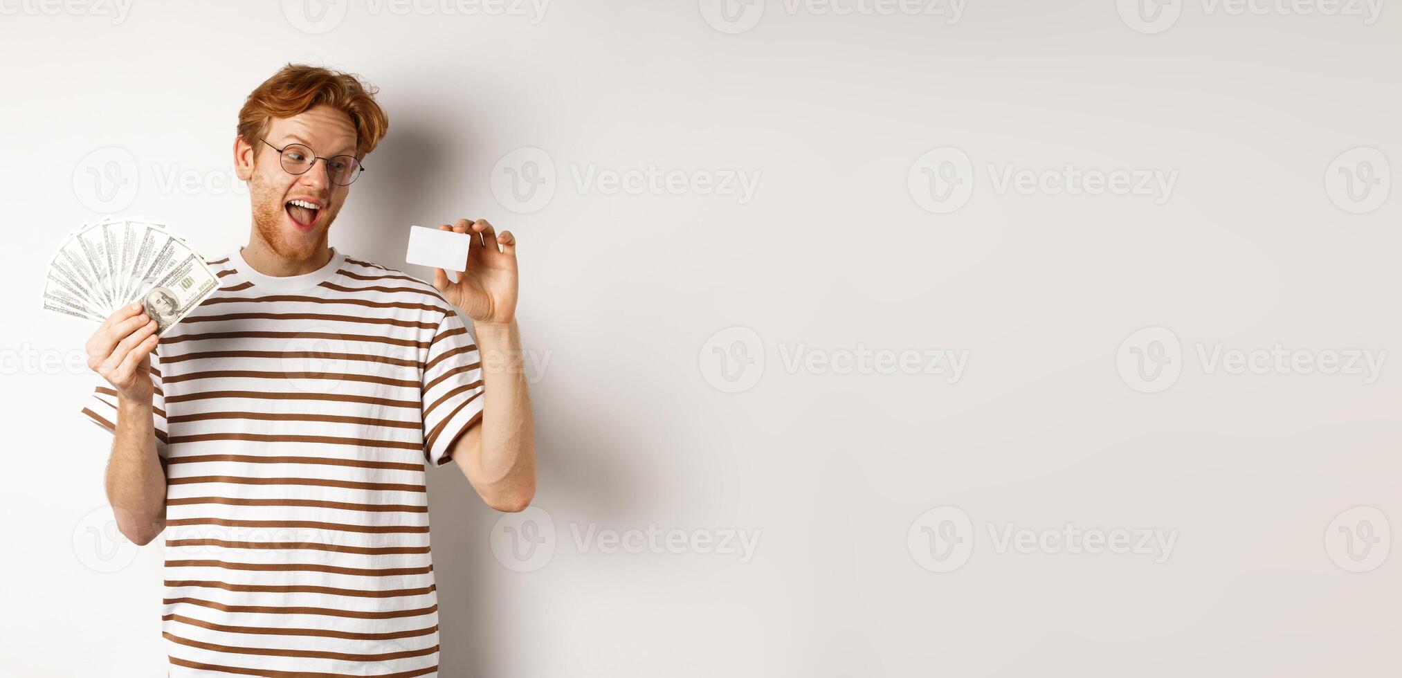 Shopping and finance concept. Young redhead man in glasses choosing plastic credit card, showing dollars and smiling, standing over white background photo