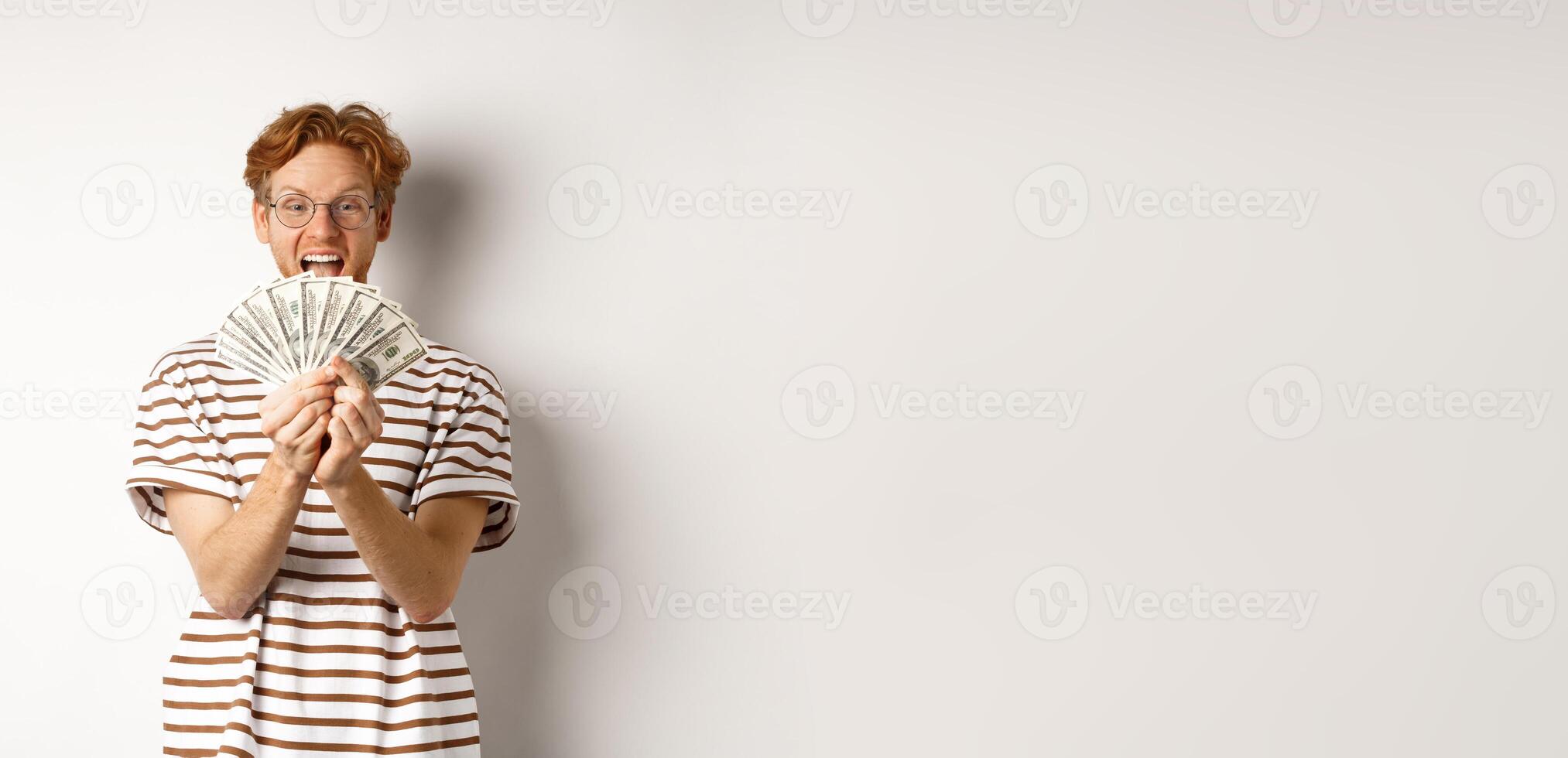 Shopping and finance concept. Lucky redhead guy winning, showing prize money and smiling happy, standing in glasses and t-shirt over white background photo