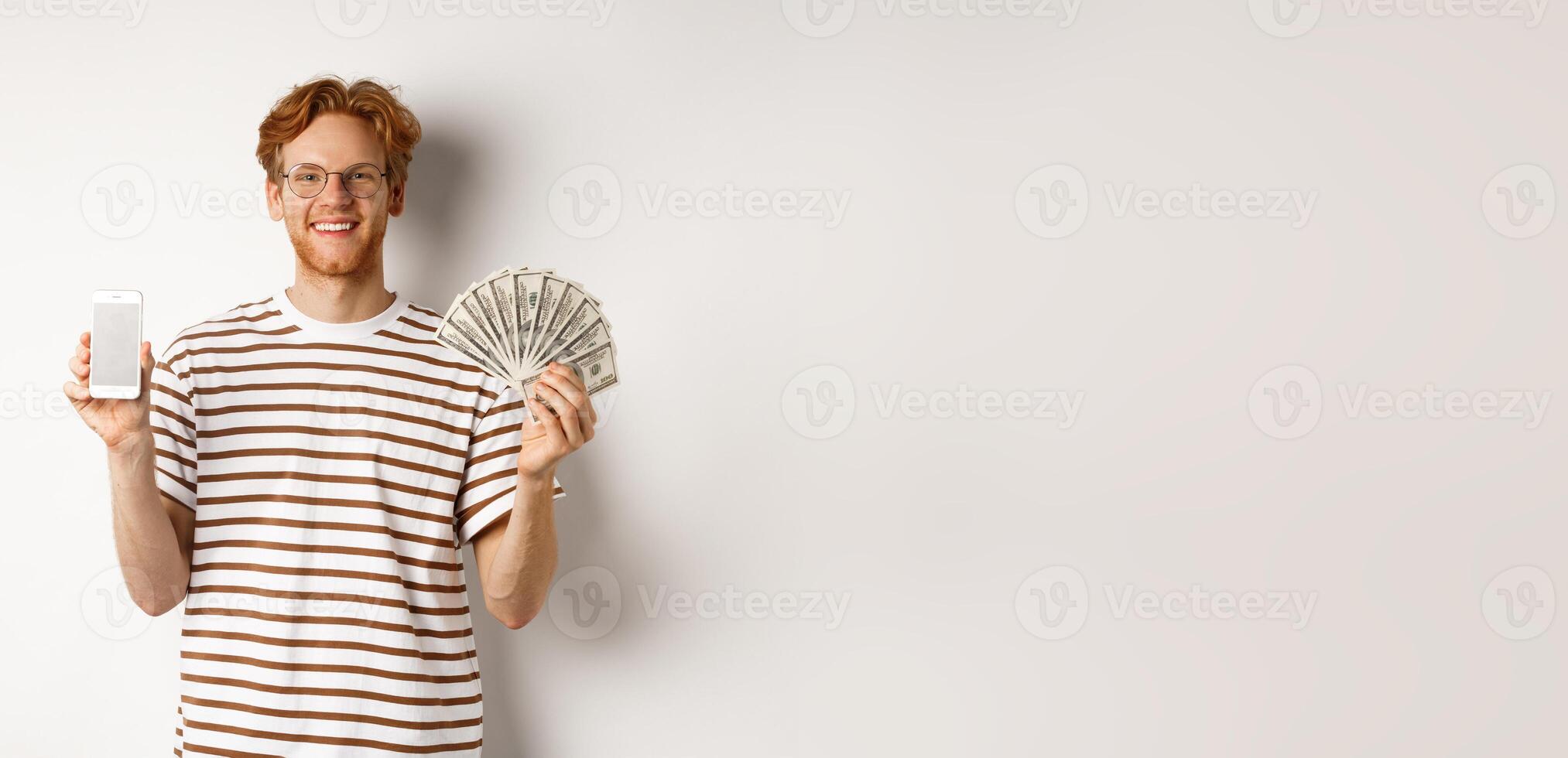 Smiling young redhead man in glasses showing smartphone blank screen and money, standing over white background photo
