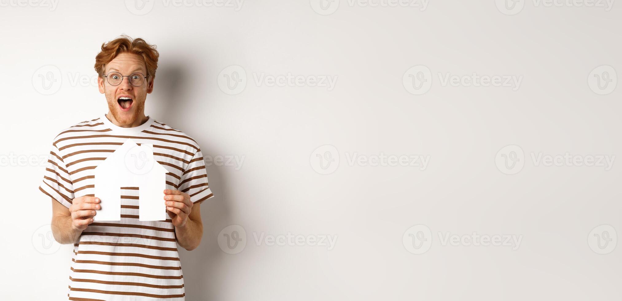 Happy and surprised redhead man winning house, holding paper home model and staring at camera, standing joyful over white background photo