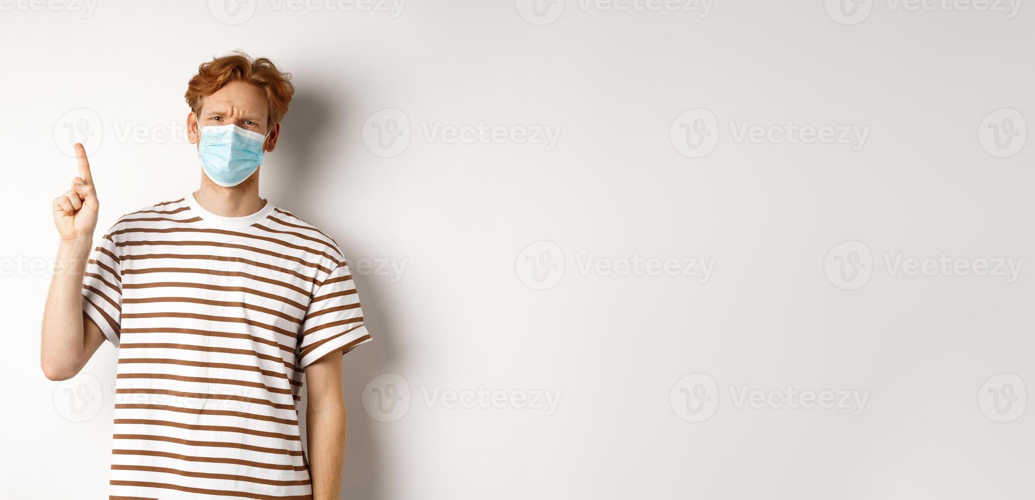 Covid, virus and social distancing concept. Disappointed young man with red hair wearing face mask, pointing finger up and frowning displeased, complaining on promo photo