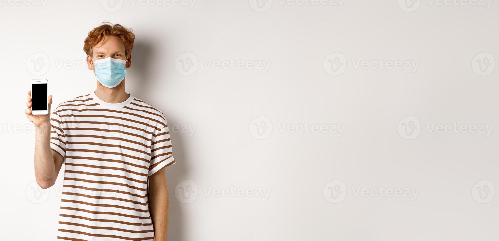 Covid-19, pandemic and social distancing concept. Handsome young man with face mask, showing black smartphone screen and smiling, standing over white background photo
