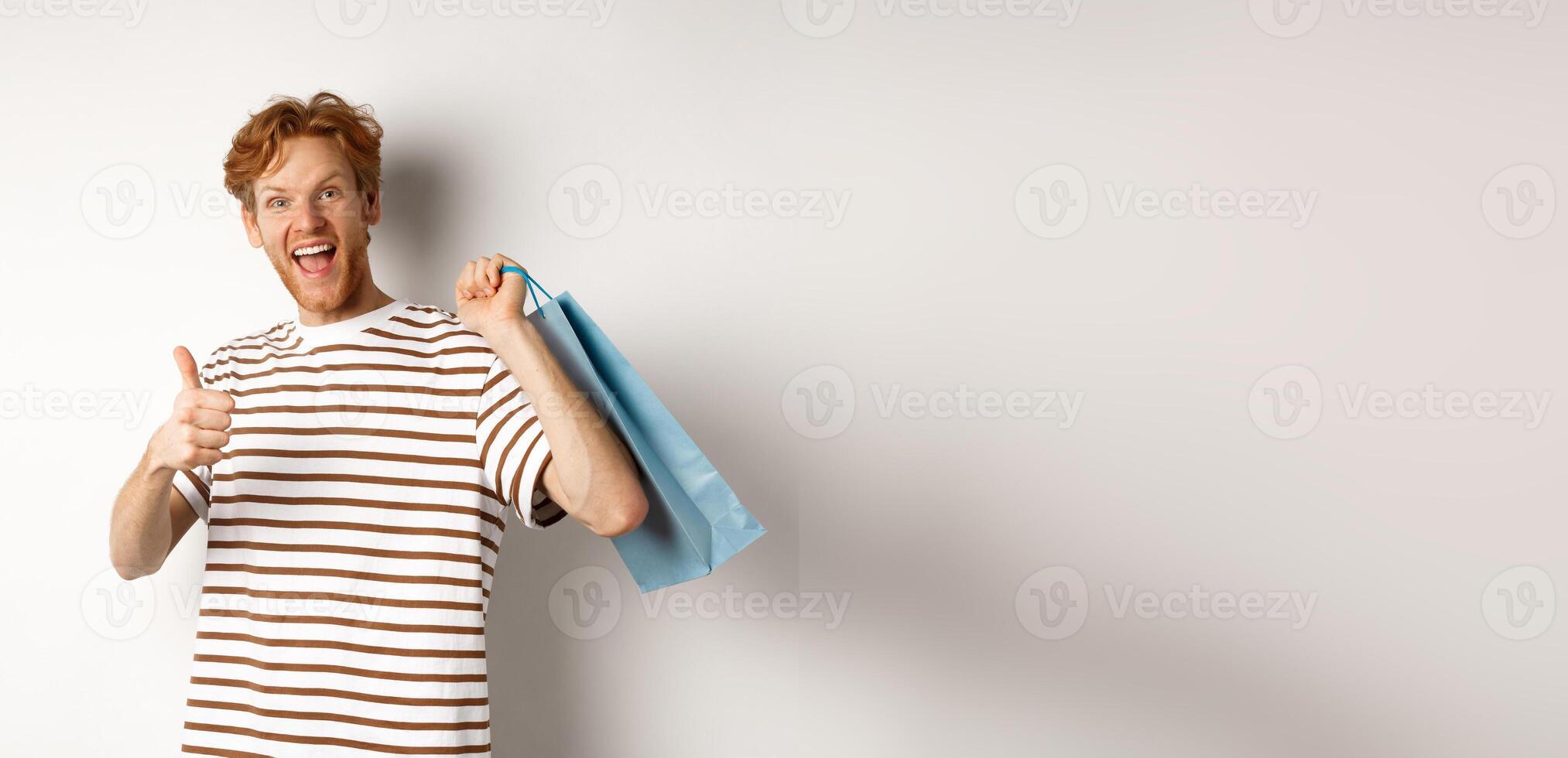 Happy young man with red hair, shopping in stores, showing thumbs-up and holding paper bag over shoulder, recommending shop, white background photo