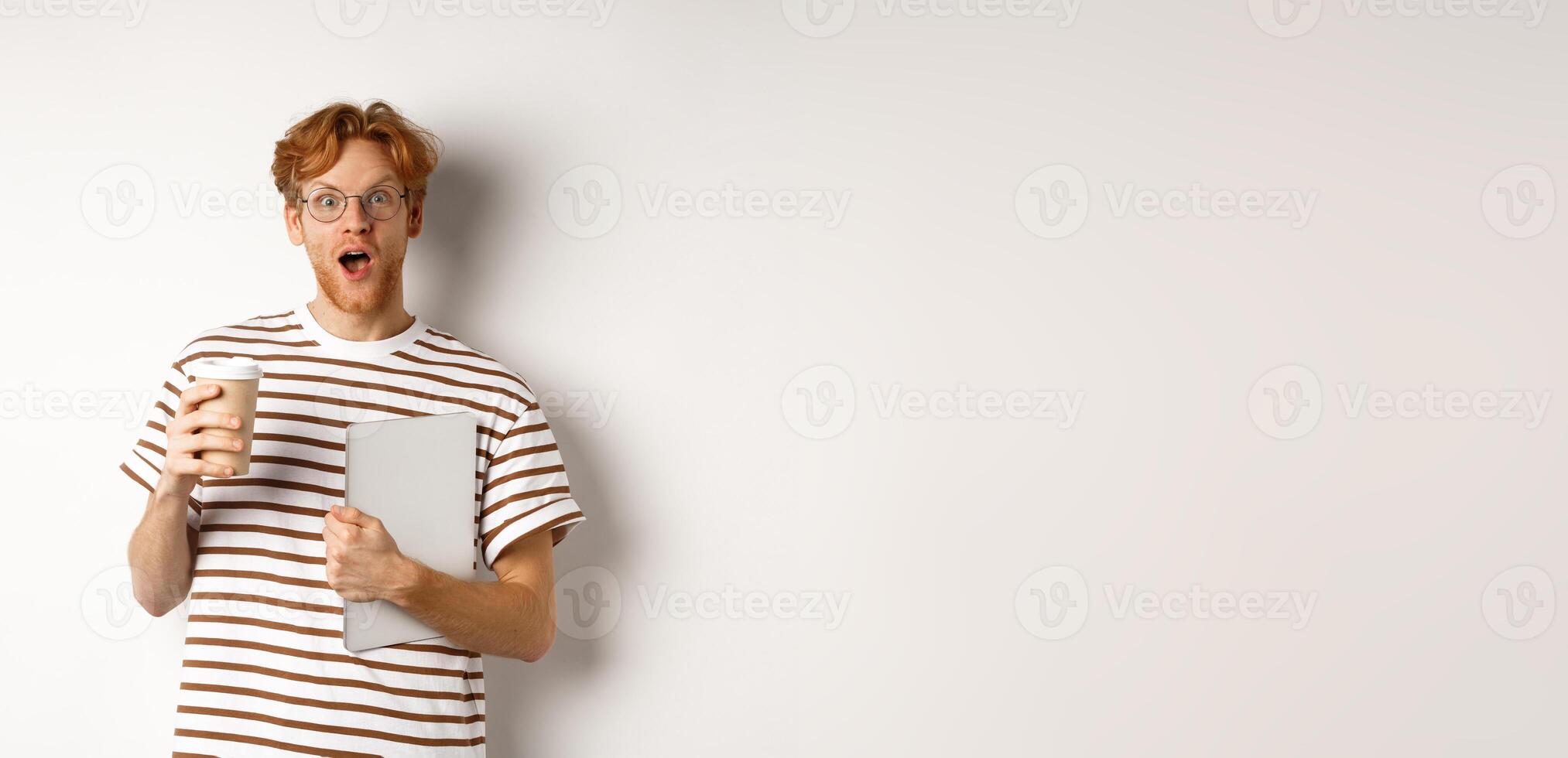 Amazed redhead man talking with coworkers on coffee break, holding cup and laptop, staring surprised at camera, white background photo