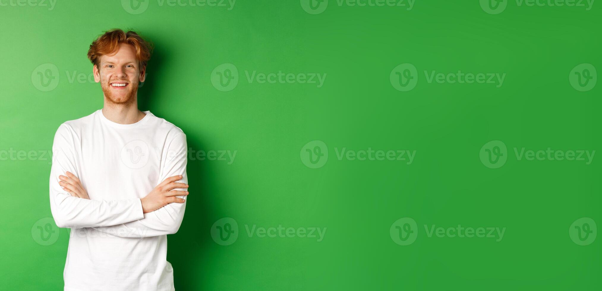 Young ambitious man with red hair standing over green background, holding hands crossed on chest and smiling photo