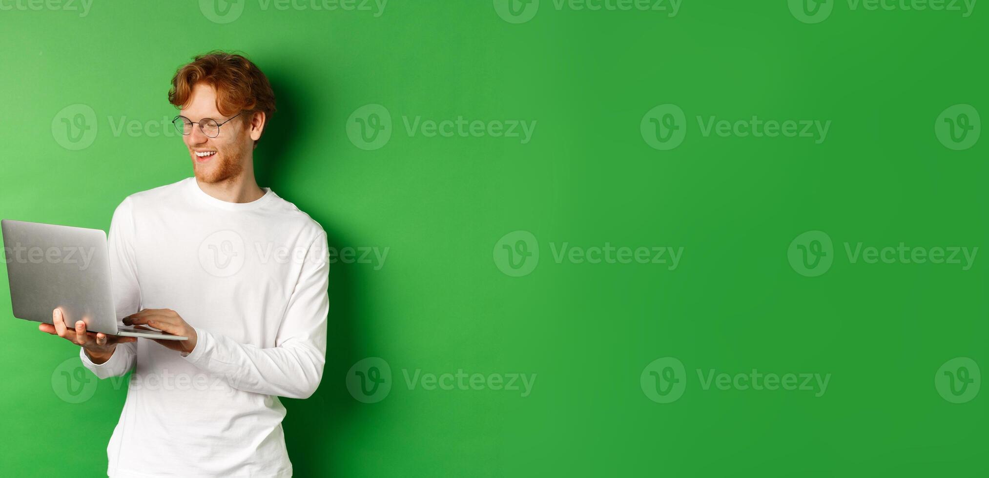 Smiling young man with red hair, wearing glasses, working on laptop and smiling, standing over green background photo