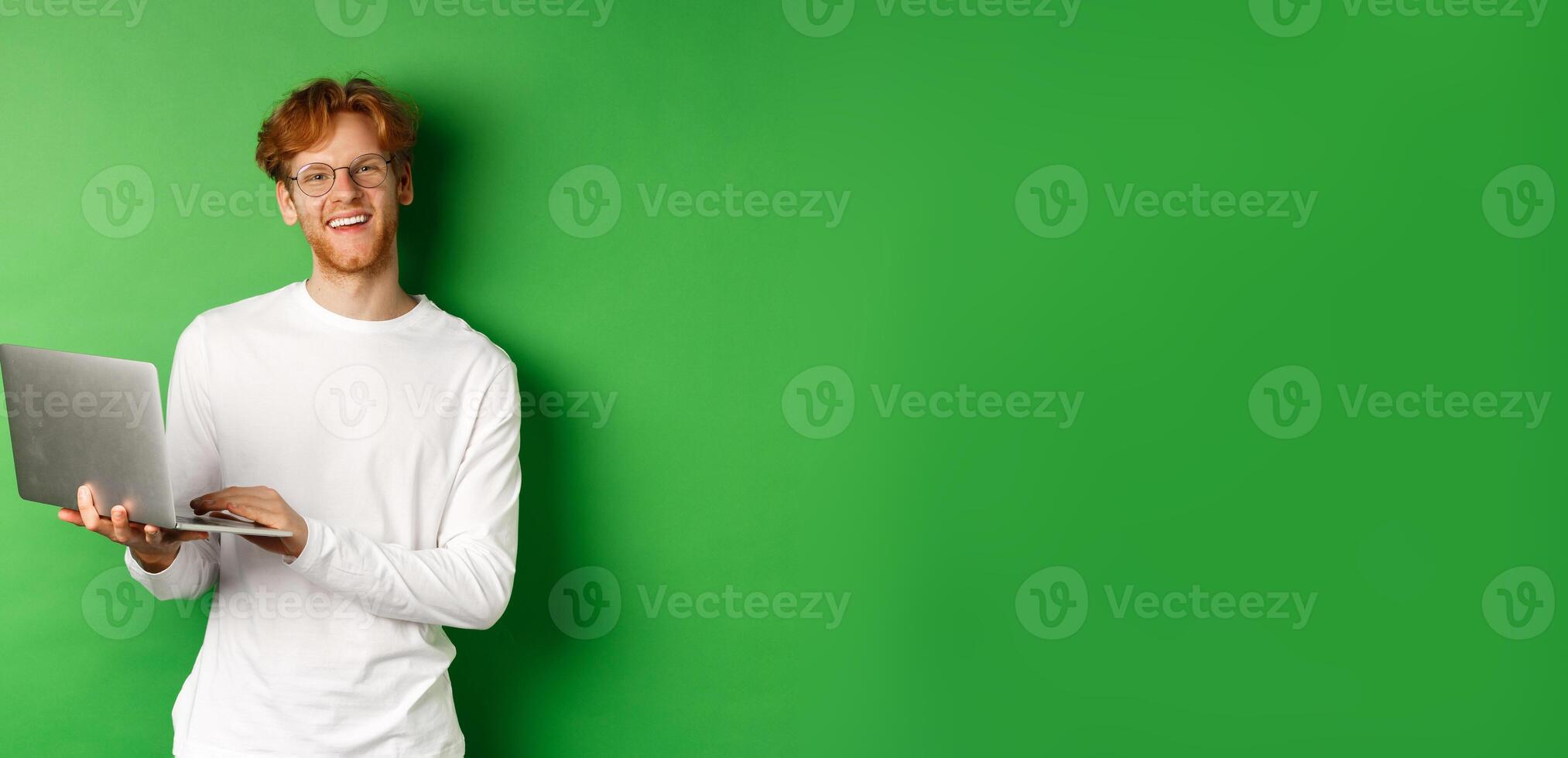 Cheerful redhead freelancer in glasses smiling at camera, working on laptop while standing over green background photo