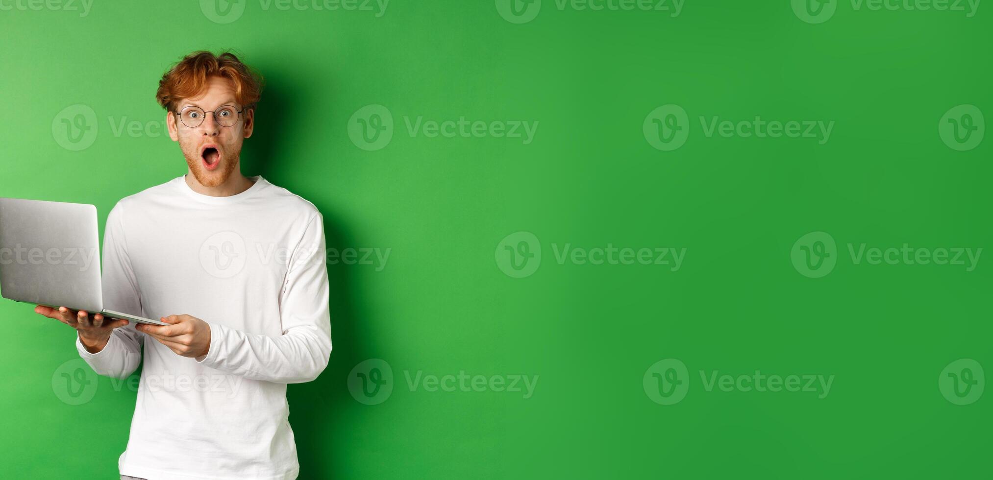 Impressed young man in glasses drop jaw and staring amazed after reading promo on laptop, standing over green background photo