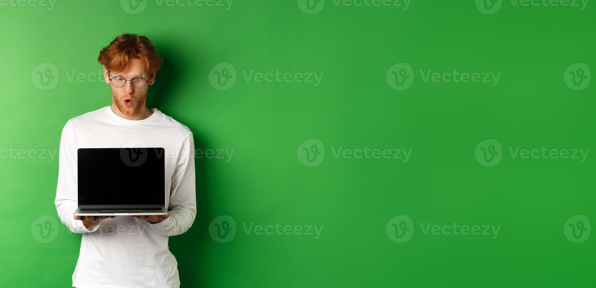 Impressed redhead nerdy guy in glasses showing blank laptop screen and looking at display amazed, standing over green background photo