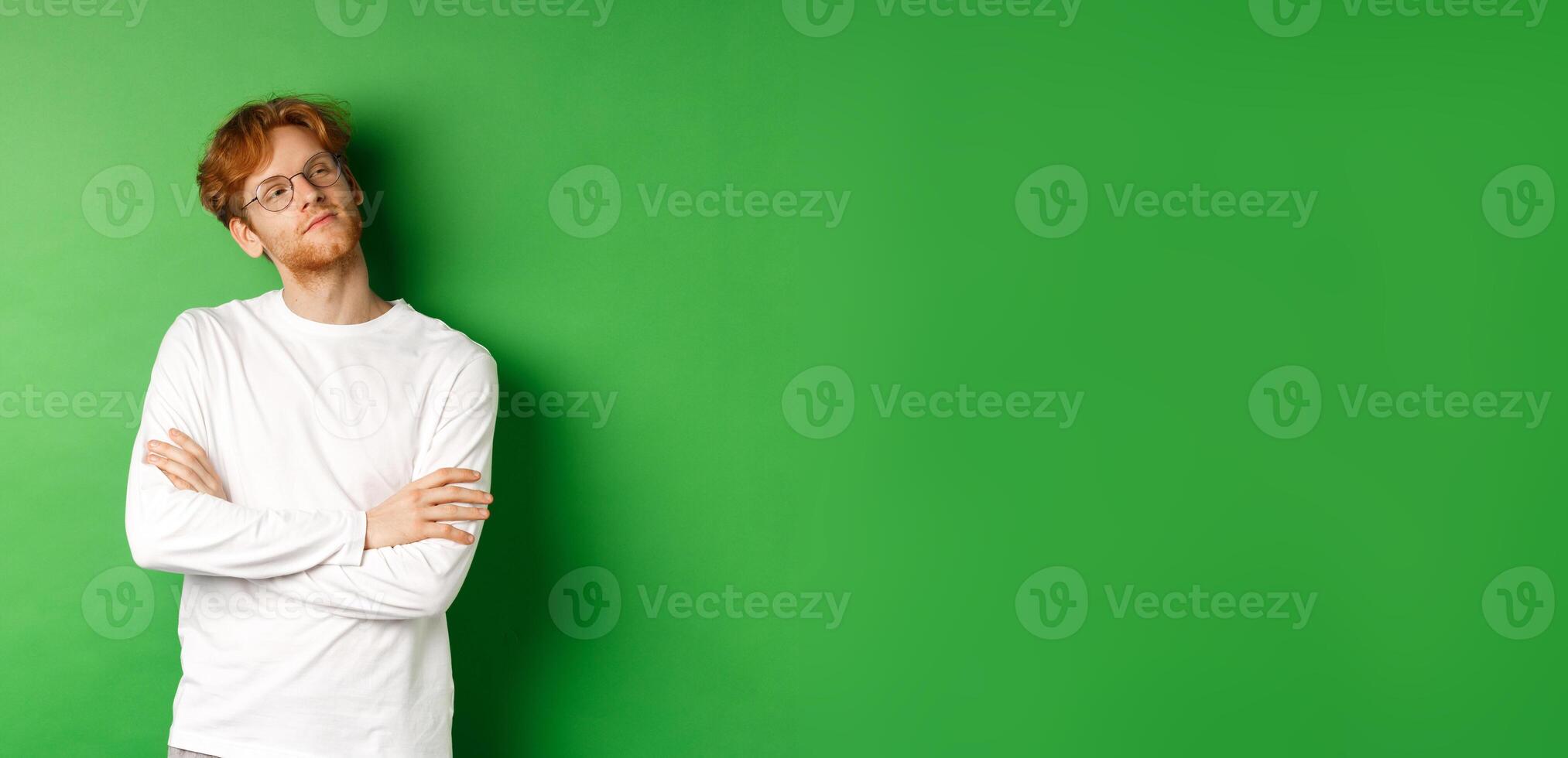 Confident young man with red hair, wearing glasses, looking smug at right side copy space, cross arms on chest self-assured, standing over green background photo