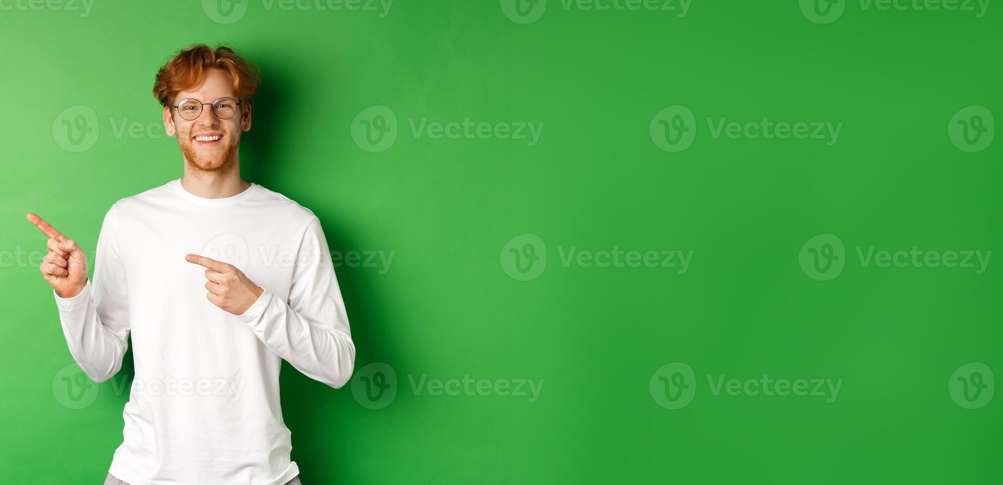 Handsome young man with red hair and beard smiling, pointing fingers left at logo, showing advertisement, standing over green background photo
