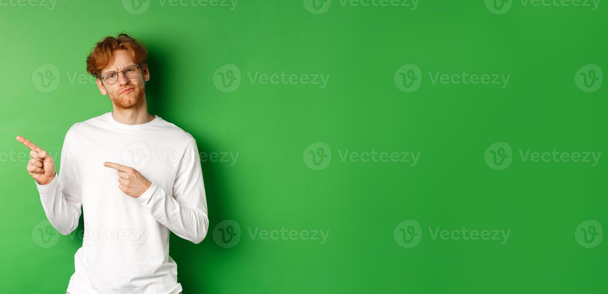 Doubtful redhead guy in glasses pointing fingers left, looking hesitant and unsure at camera, standing over green background photo