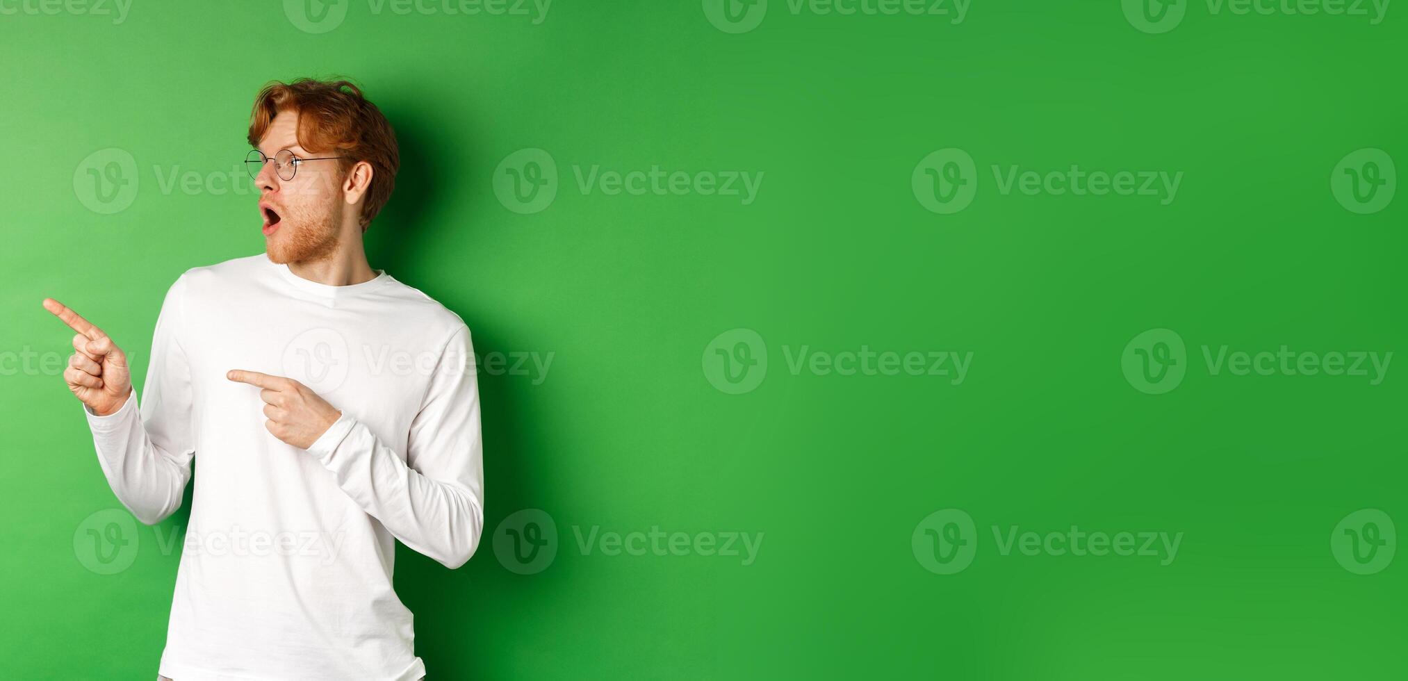 Impressed redhead man in glasses checking out promo, pointing and looking left with amazed face, standing in long-sleeve t-shirt over green background photo