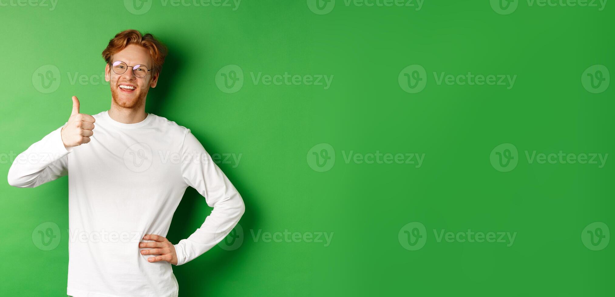 Cheerful young man with beard and red hair, wearing glasses, showing thumb-up in approval and smiling, say yes, standing over green background photo