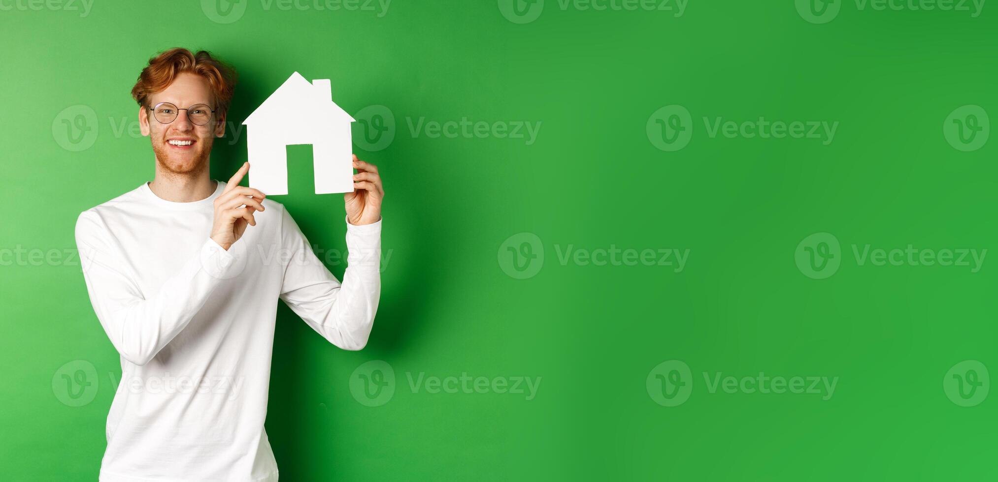 Real estate. Handsome young man with red hair, wearing glasses, showing paper house cutout and smiling, standing against green background photo