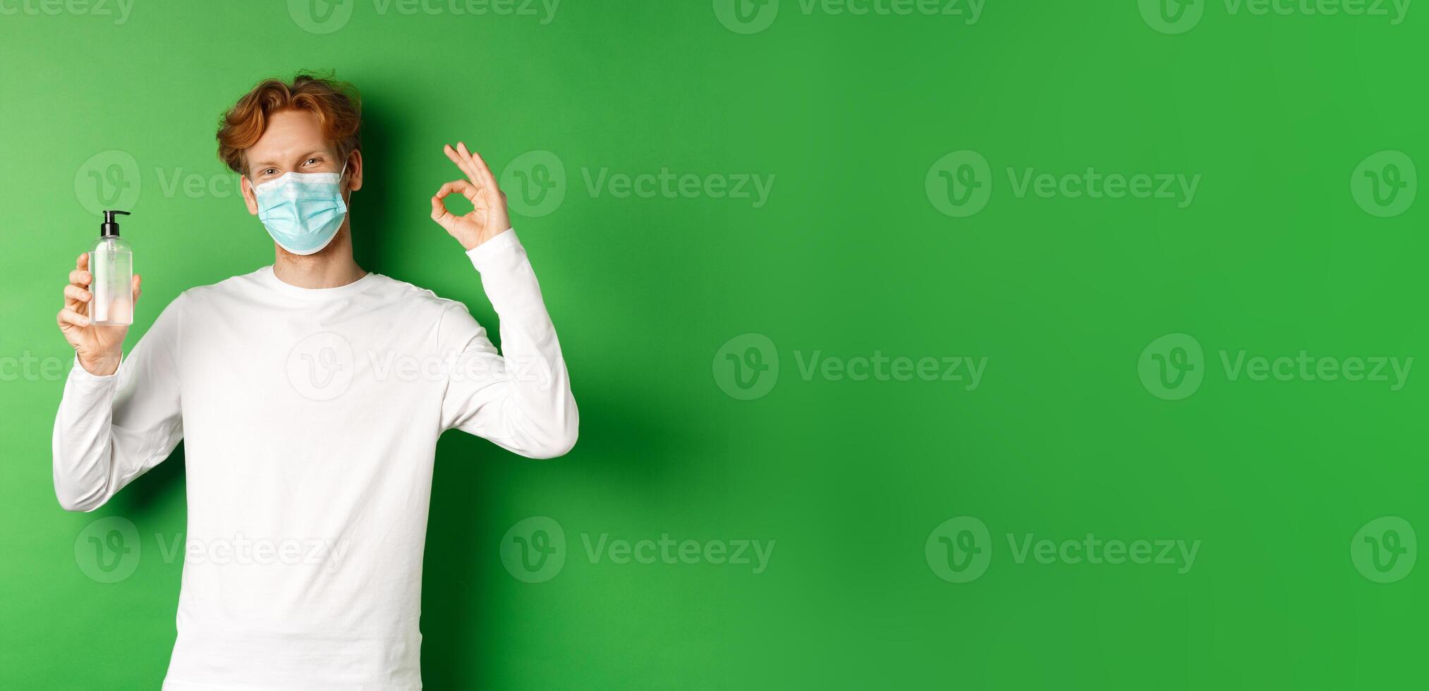 Covid-19, virus and social distancing concept. Smiling young man with red hair, wearing face mask from coronavirus, showing okay sign and hand sanitizer, green background photo