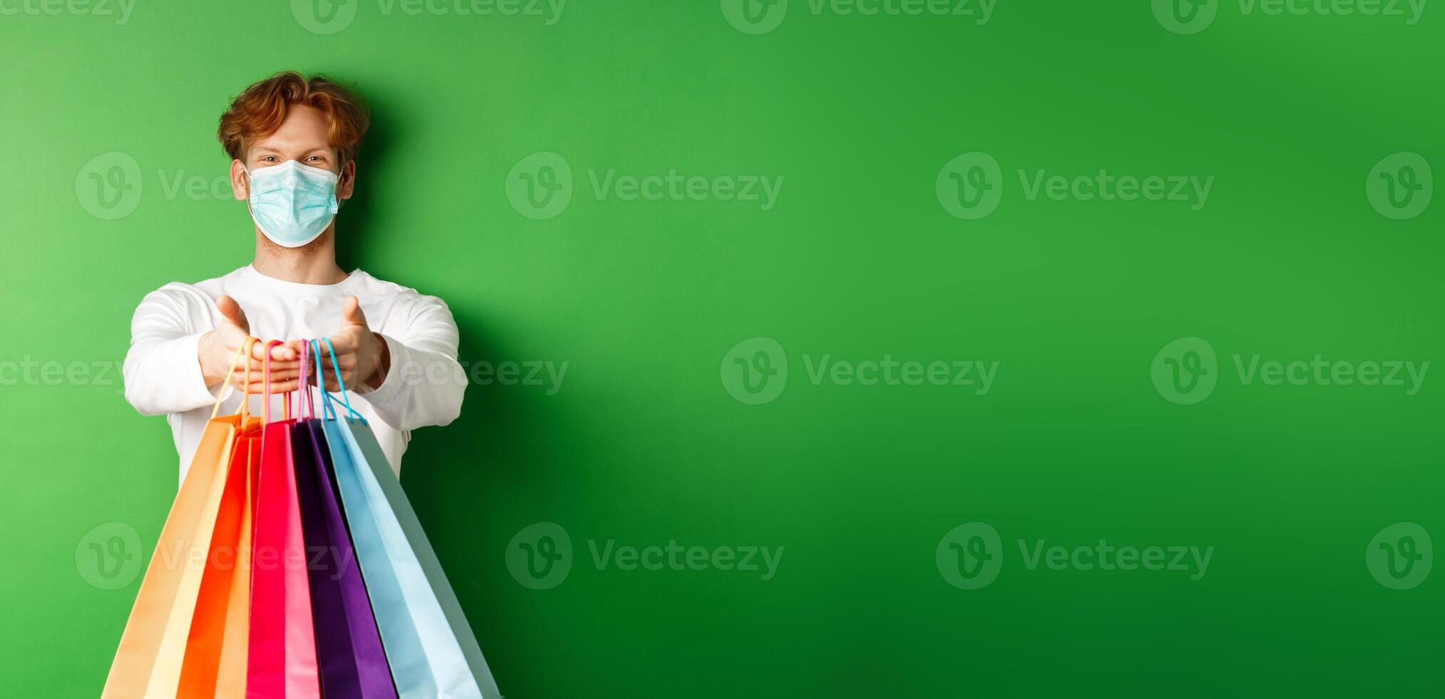 Happy young man in medical mask giving you shopping bags with purchases, smiling and wishing well, standing over green background. Covid-19 concept photo
