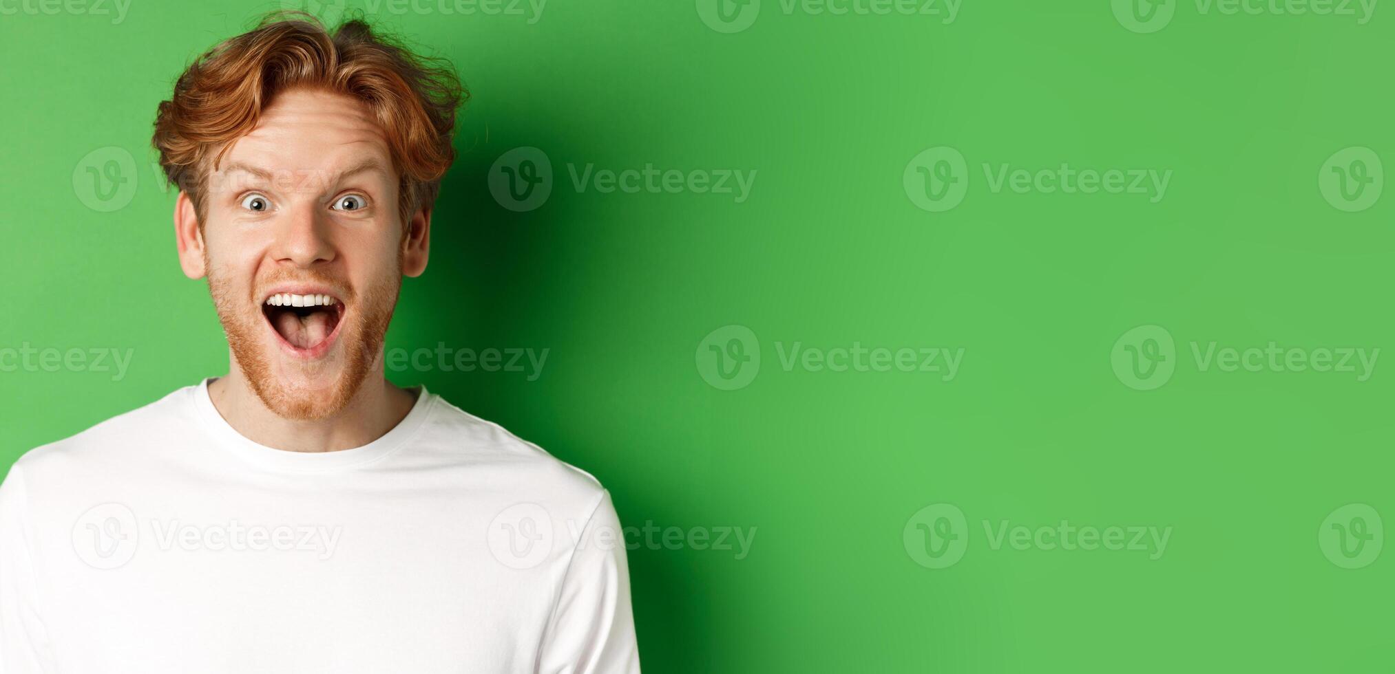 Emotions and fashion concept. Close up of surprised redhead man hear amazing news, staring at camera joyful, standing over green background photo