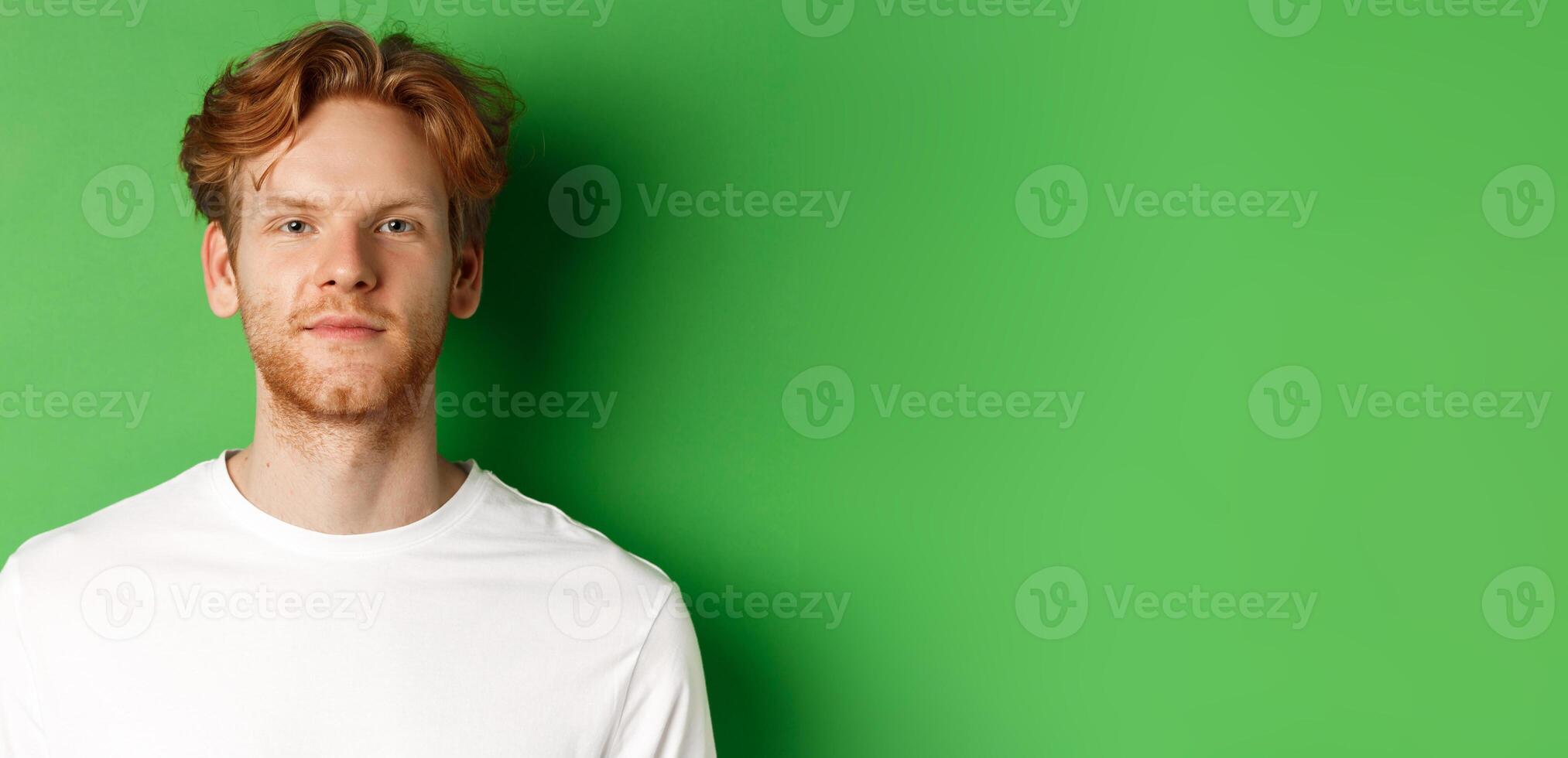 Close-up of young man with red messy hair and beard looking at camera, standing over green background photo