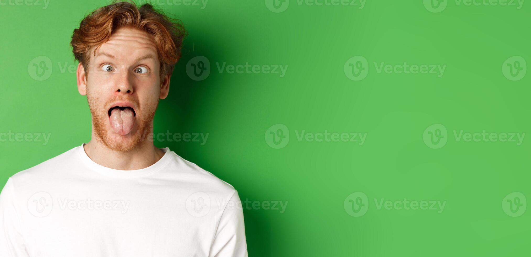 Emotions and fashion concept. Close up of funny redhead man showing silly faces, sticking tongue and squinting, standing over green background photo