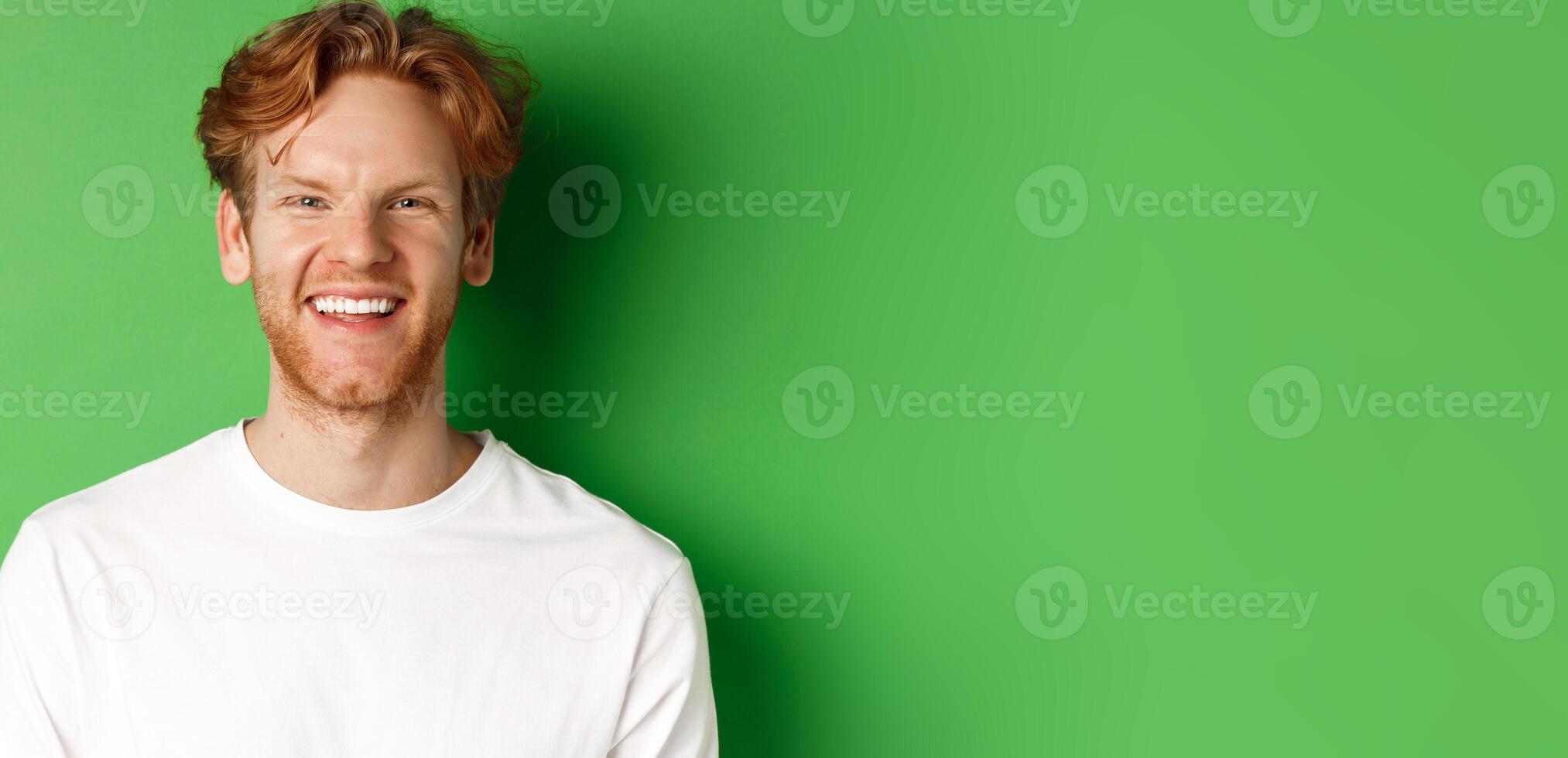 Emotions and fashion concept. Happy young man with red hair and beard, smiling and laughing at camera, standing over green background photo