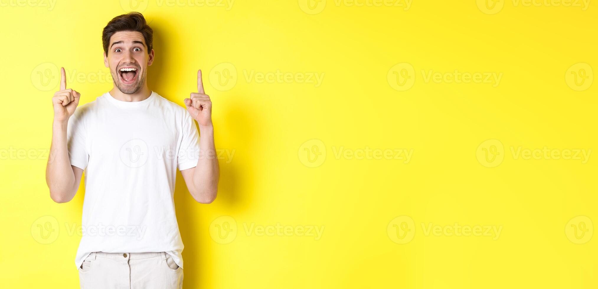 Portrait of excited handsome man in white t-shirt, pointing fingers up, showing offer, standing against yellow background photo