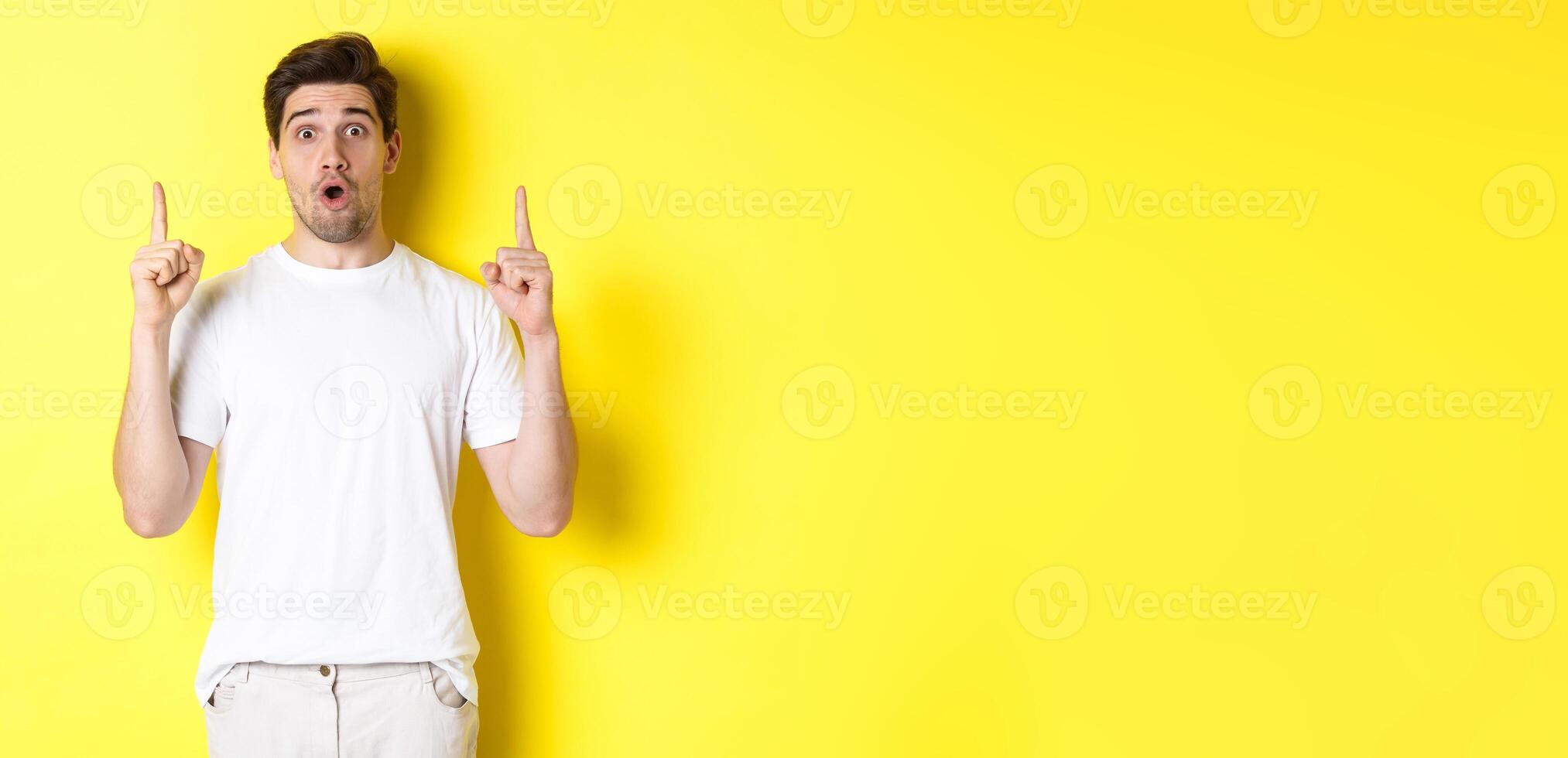 Surprised handsome guy in white t-shirt, pointing fingers up, interested about advertisement, standing against yellow background photo