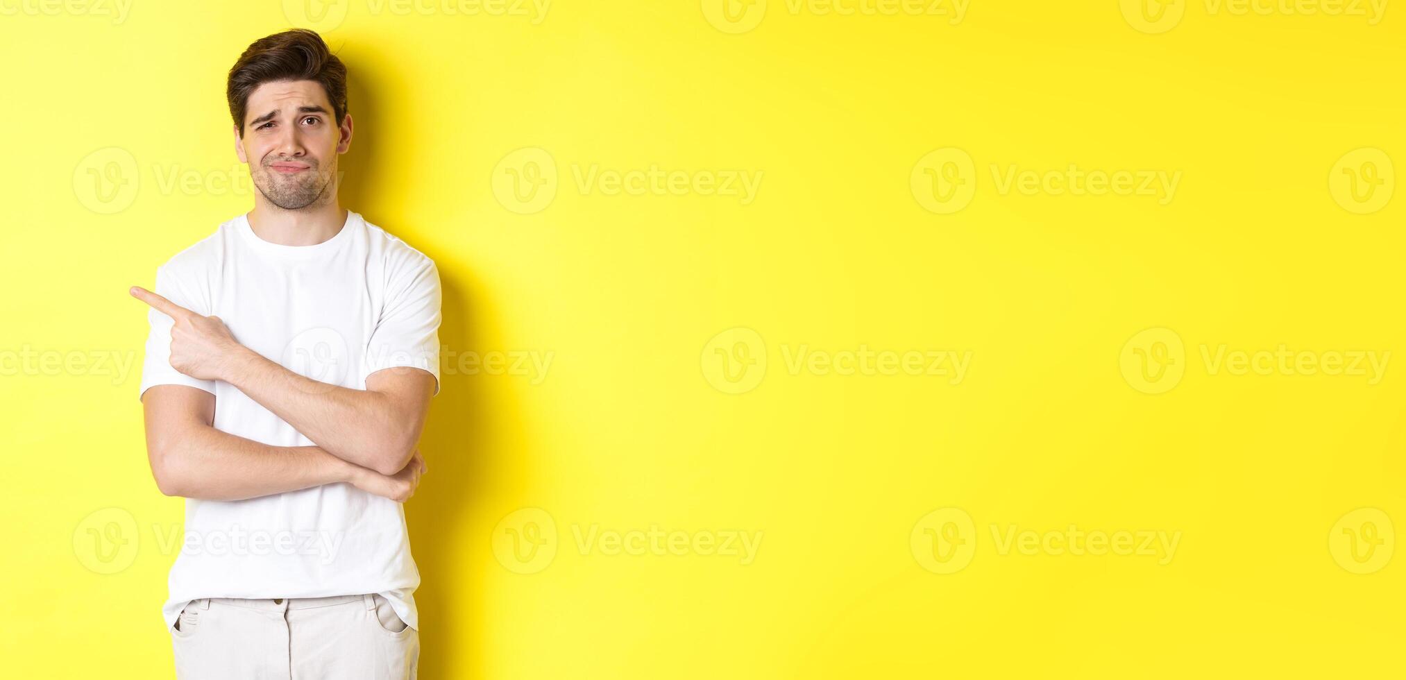 Skeptical and reluctant guy in white t-shirt, pointing finger left and smirking unamused, showing bad advertisement, standing over yellow background photo