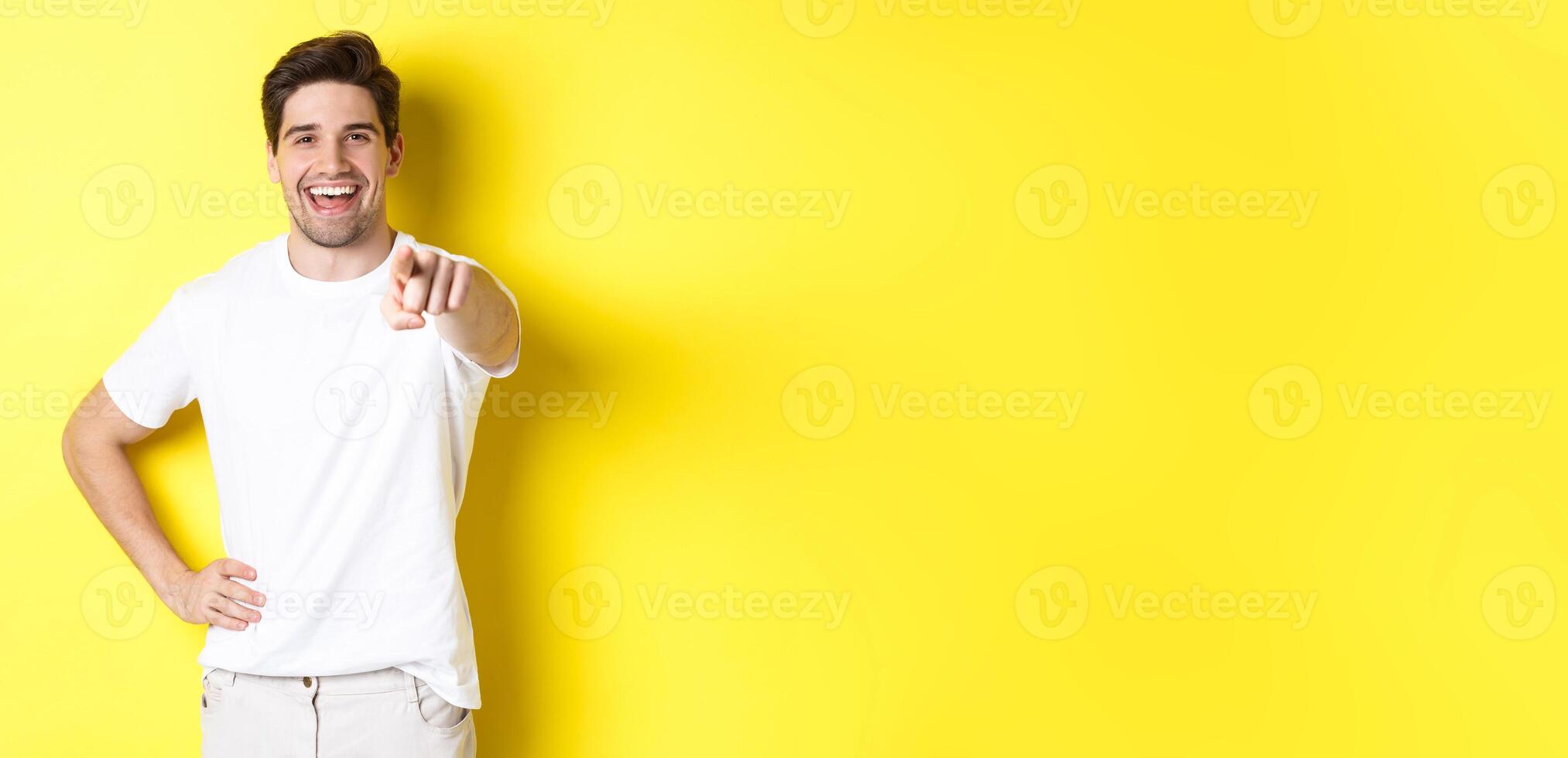 Confident smiling man pointing at you camera, standing in white clothes against yellow background photo