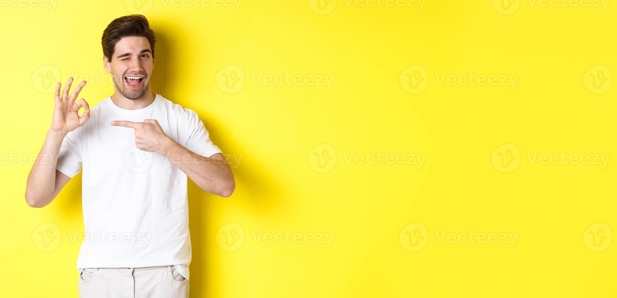 Image of handsome young man approve something, showing okay sign and winking, standing against yellow background photo