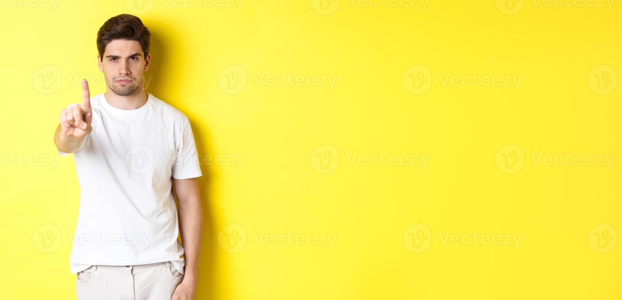 Confident man saying no, decline and prohibit something, showing one finger and frowning, standing over yellow background photo
