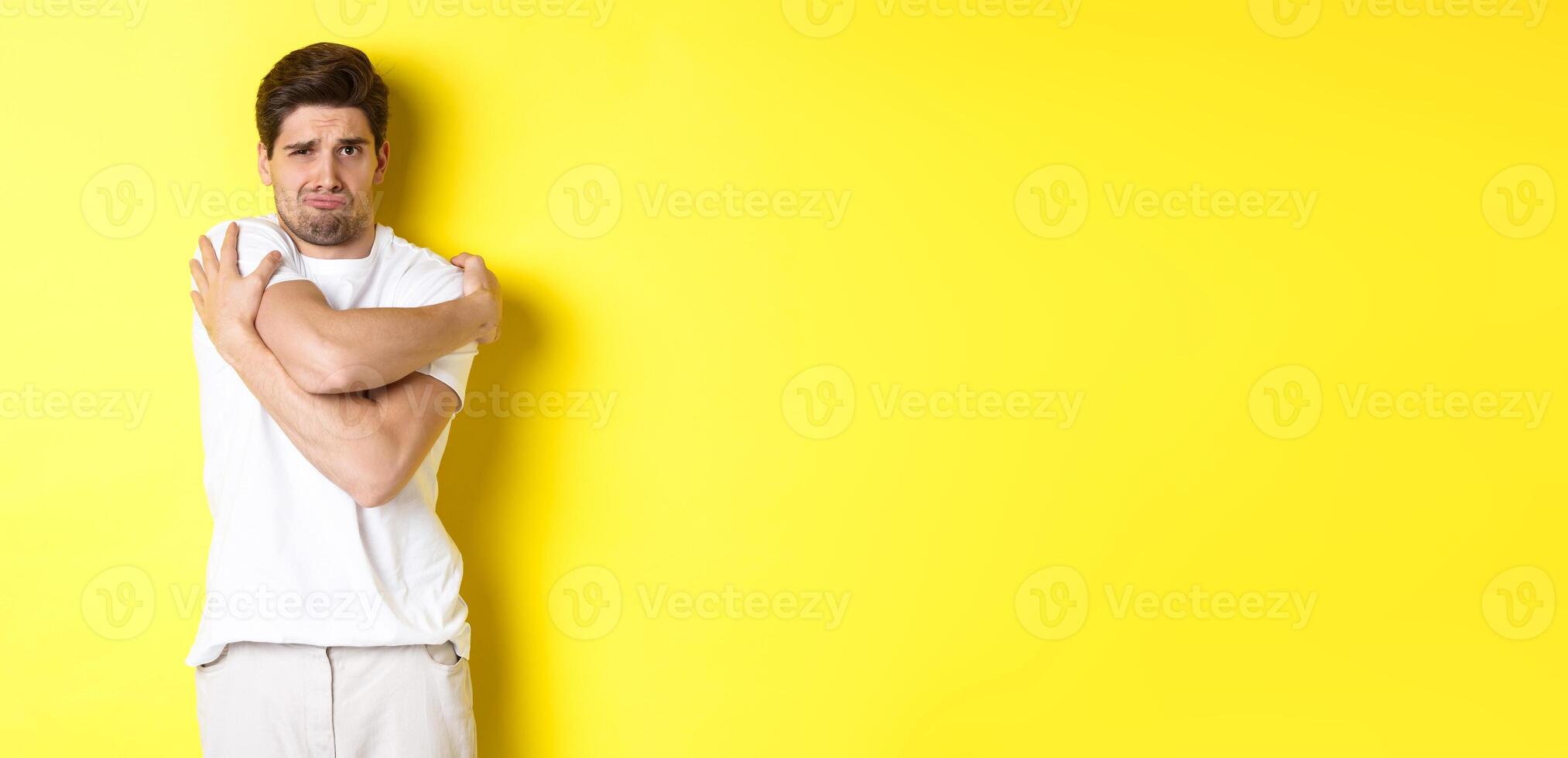 Silly and timid guy trying to comfort himself, hugging his body and frowning scared, standing over yellow background photo