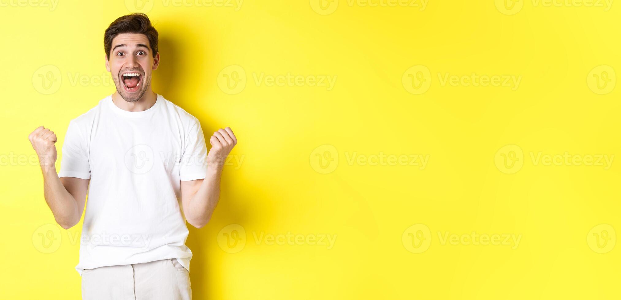 Excited and lucky man winning, clench fists and looking happy, triumphing and celebrating, standing over yellow background photo