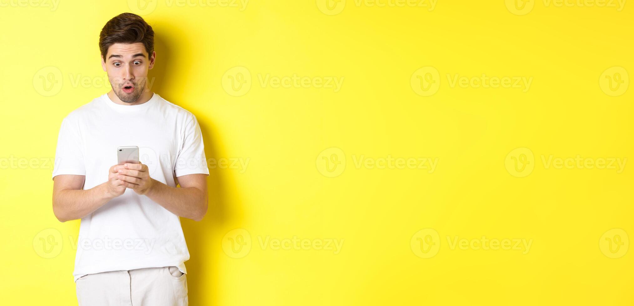Man looking surprised in smartphone, reading message on cell phone, standing in white outfit against yellow background photo