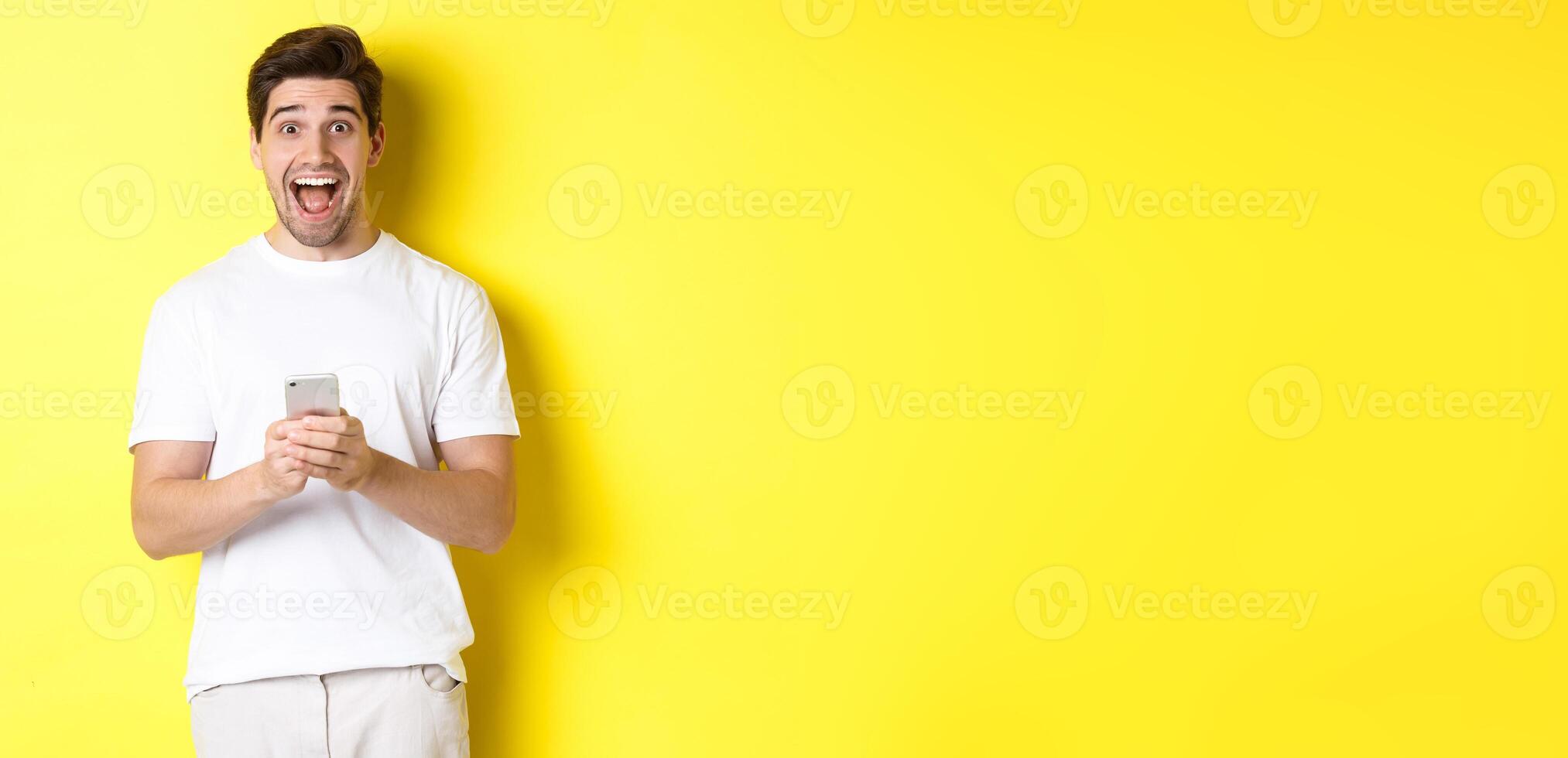 Man looking amazed and happy after reading something on mobile phone, standing over yellow background photo