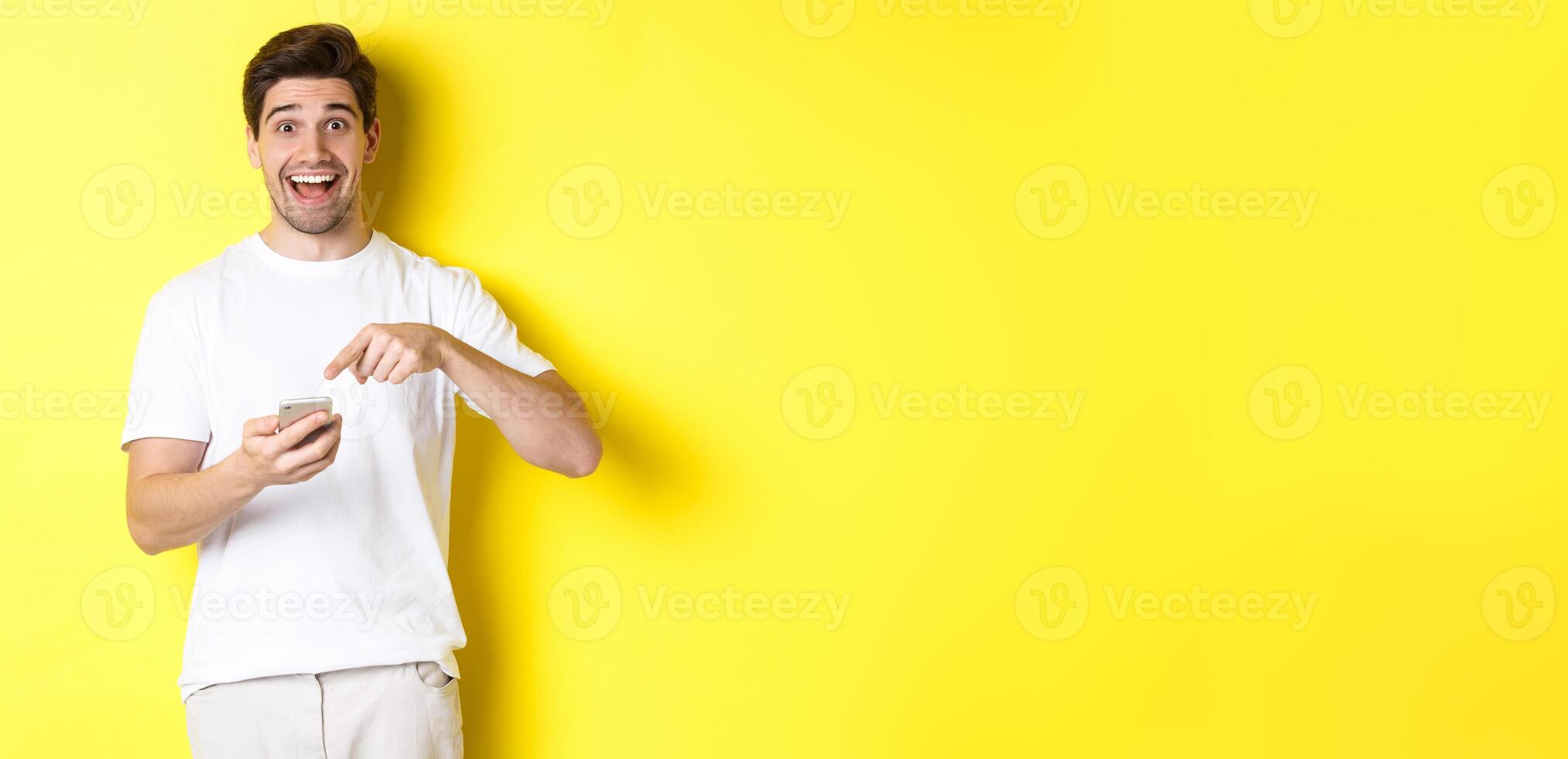 Happy man pointing at smartphone to show promo, check out internet offer, standing over yellow background photo