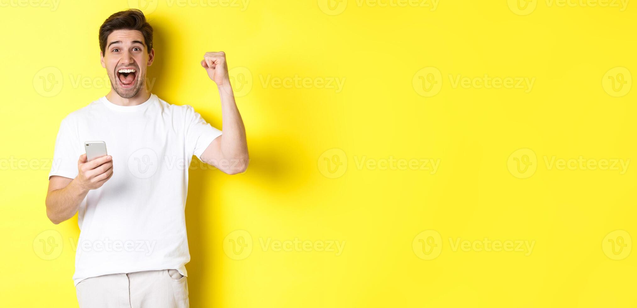 Cheerful man winning on smartphone, raising hand up and holding mobile, achieve app goal, standing over yellow background photo