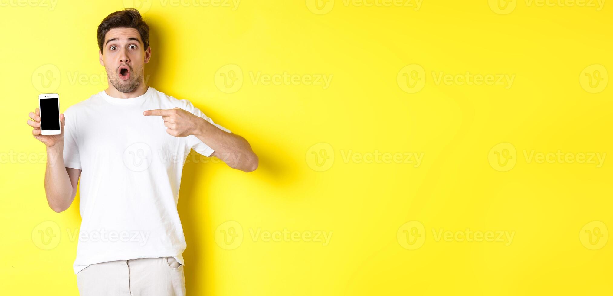 Impressed guy check out smartphone screen, pointing finger at display and gasping amazed, standing over yellow background photo
