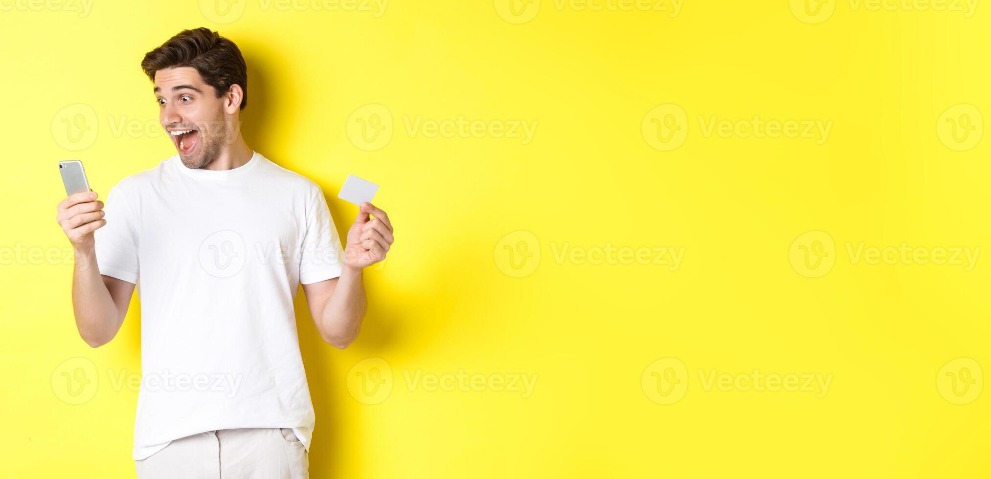 Surprised guy holding smartphone and credit card, online shopping on black friday, standing over yellow background photo