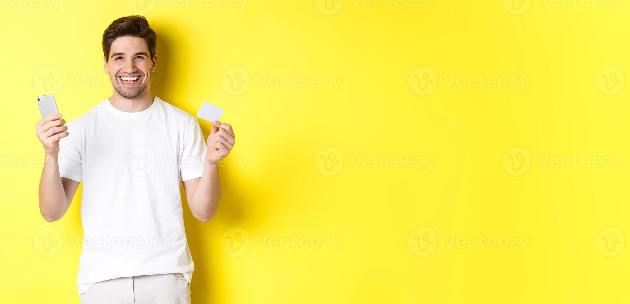 Happy young man shopping online in smartphone, holding credit card and smiling, standing over yellow background photo