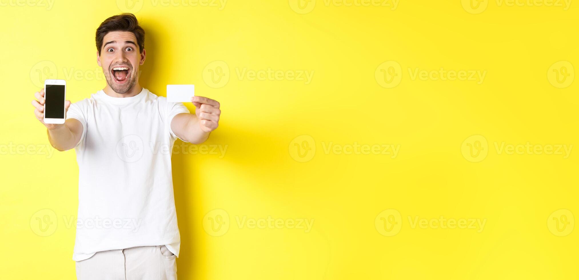 Handsome caucasian man showing smartphone screen and credit card, concept of mobile banking and online shopping, yellow background photo