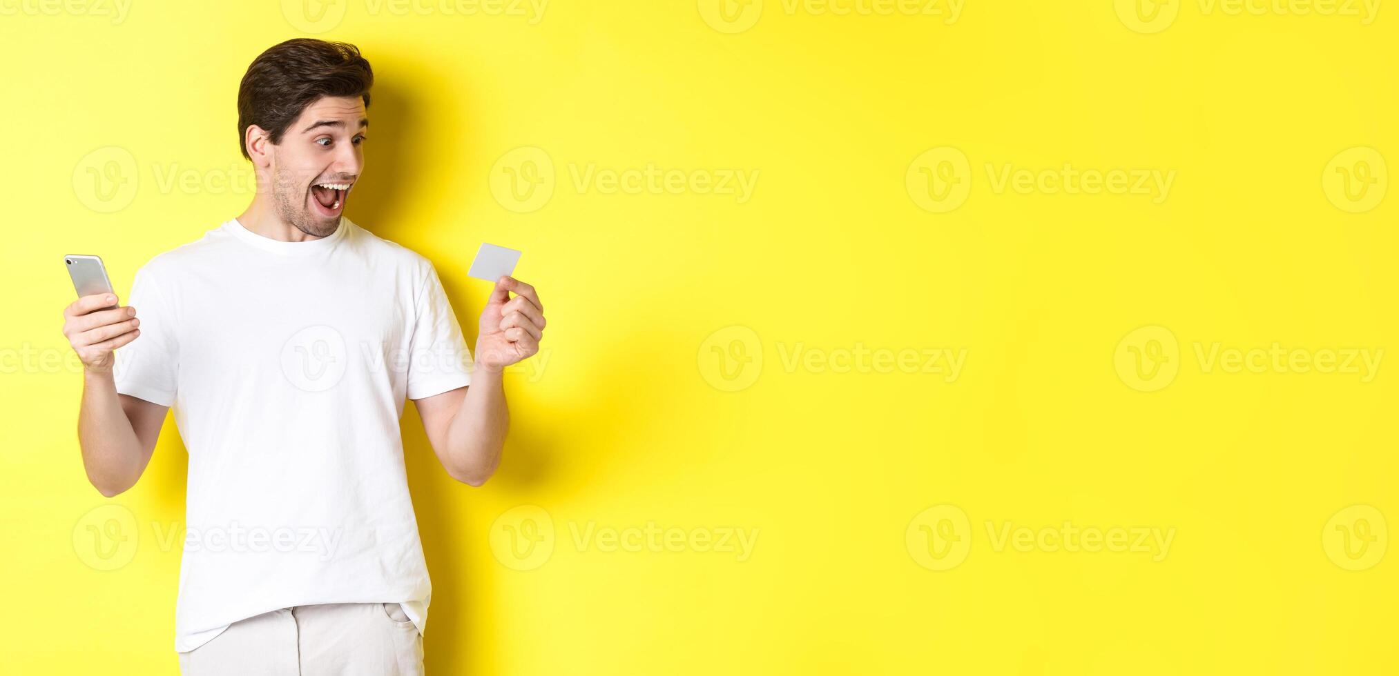 Surprised guy holding smartphone and credit card, online shopping on black friday, standing over yellow background photo