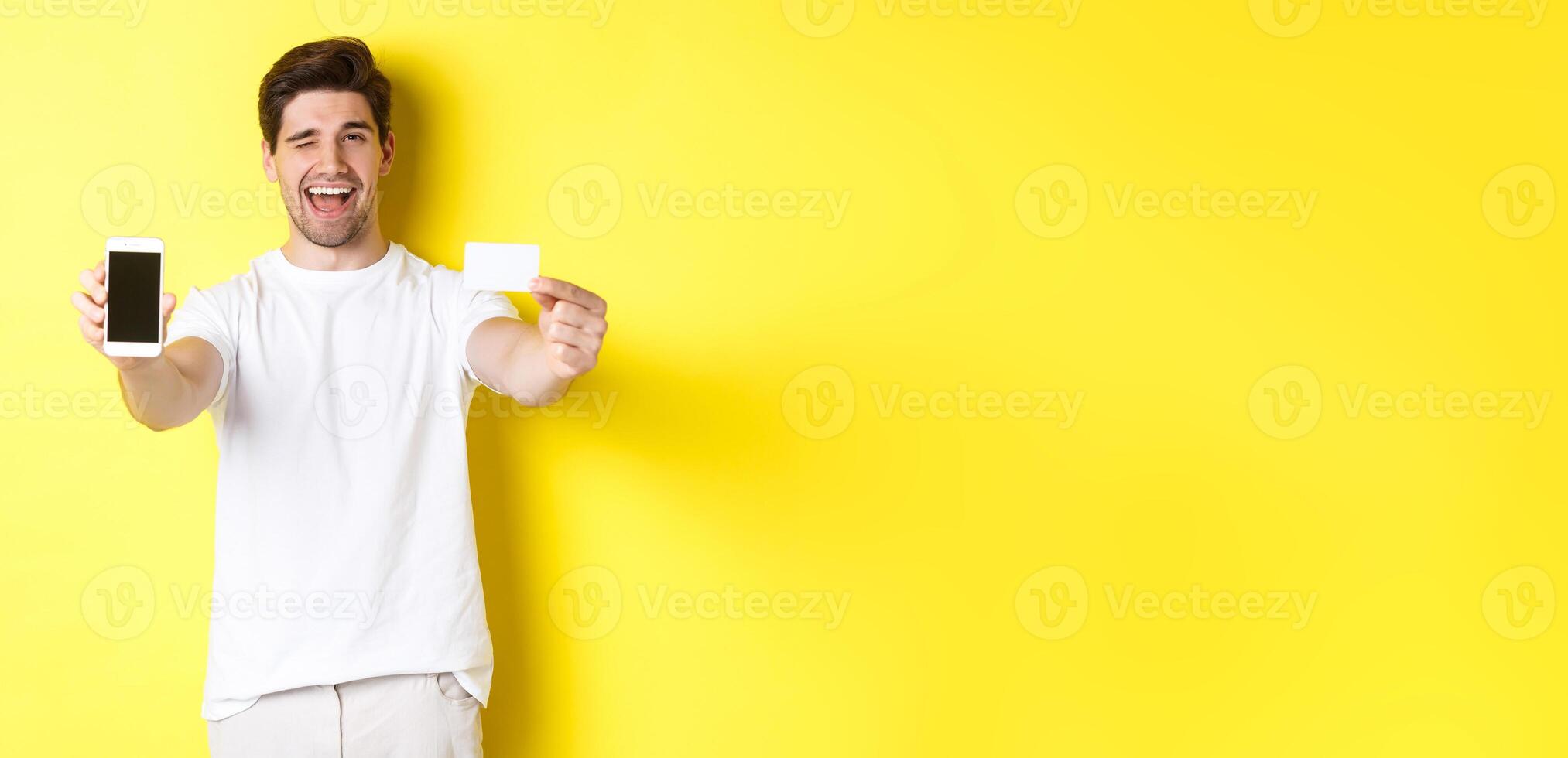 Happy man showing good online offer on mobile phone screen, holding credit card and winking, standing over yellow background photo