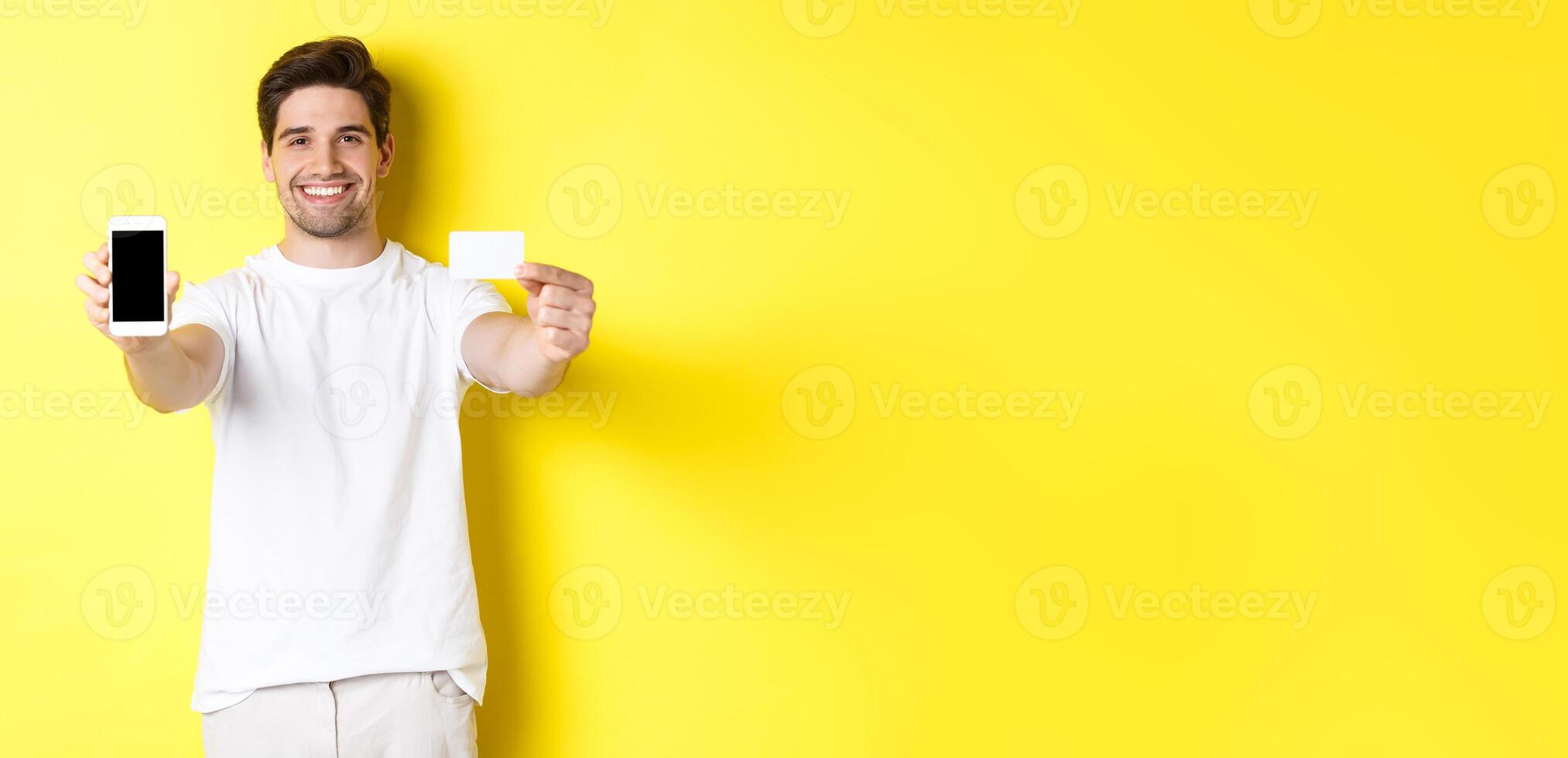 Handsome caucasian male model showing smartphone screen and credit card, concept of mobile banking and online shopping, yellow background photo