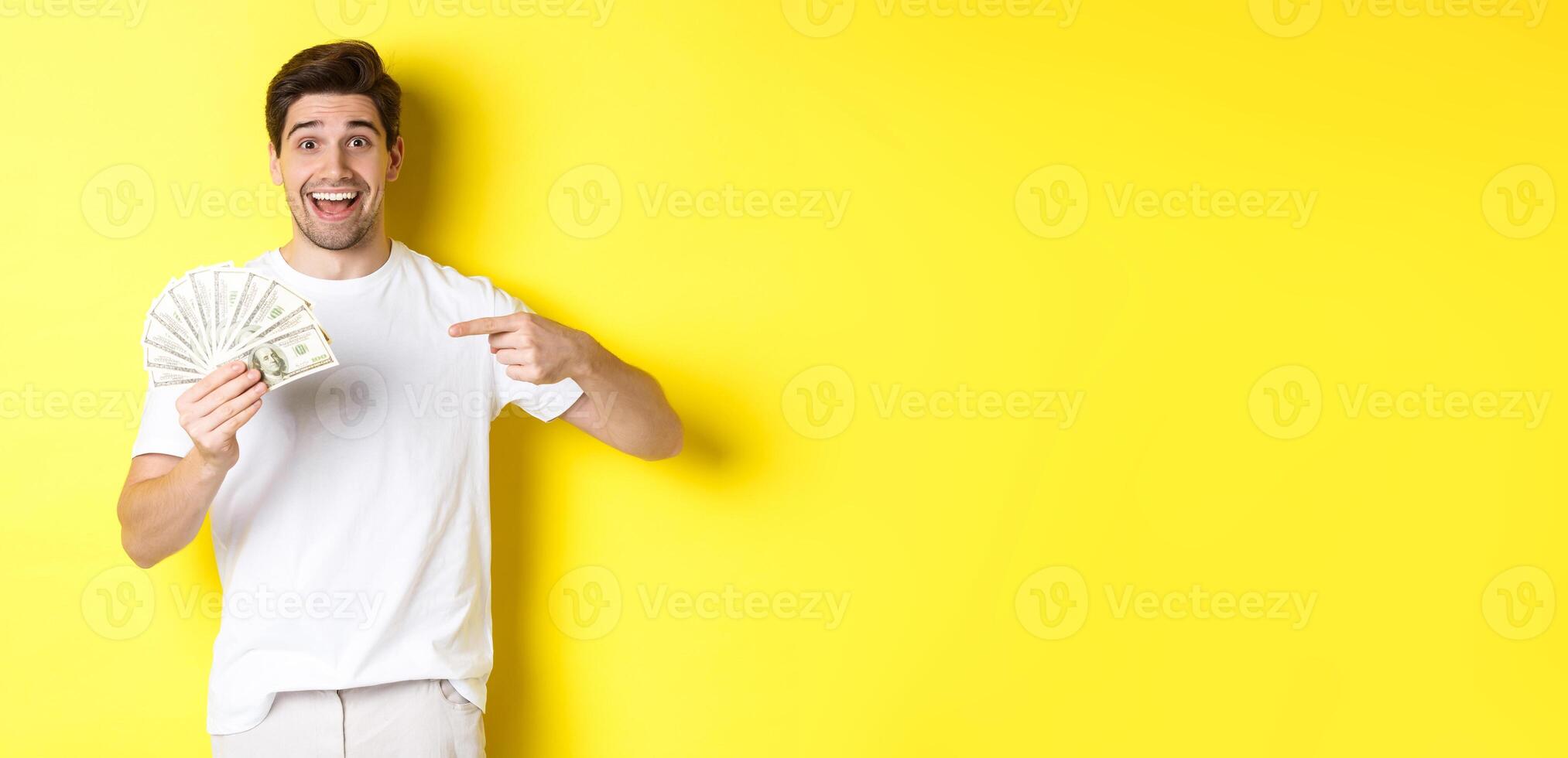 Happy handsome guy pointing finger at money, concept of credit and loan, standing over yellow background photo